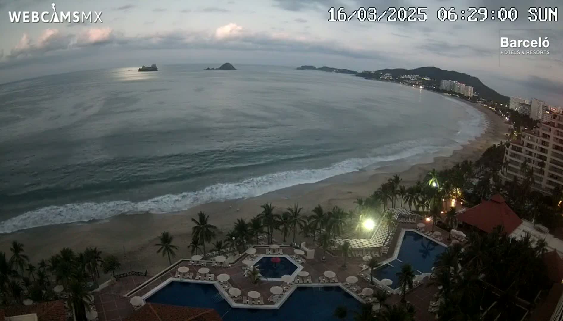 An ocean beach with moderate waves under a cloudy sky is viewed from above a resort with multiple swimming pools.