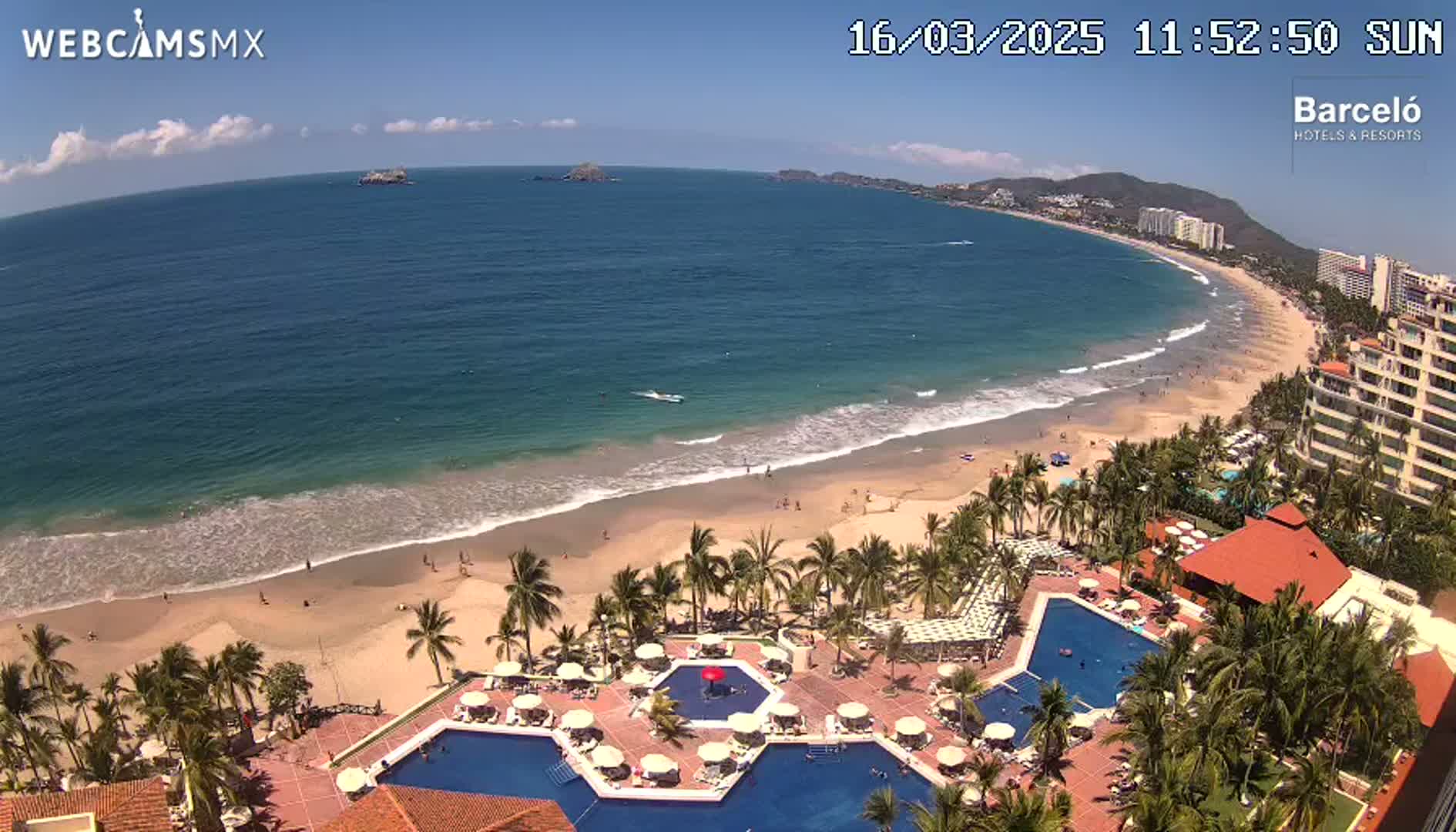 An aerial view shows a resort with multiple pools overlooking a sandy beach and ocean on a sunny day with some clouds.