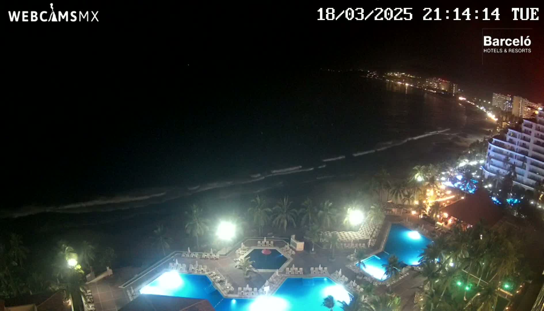 A nighttime aerial view shows a resort with illuminated pools and palm trees next to a dark ocean with waves under a clear, dark sky.
