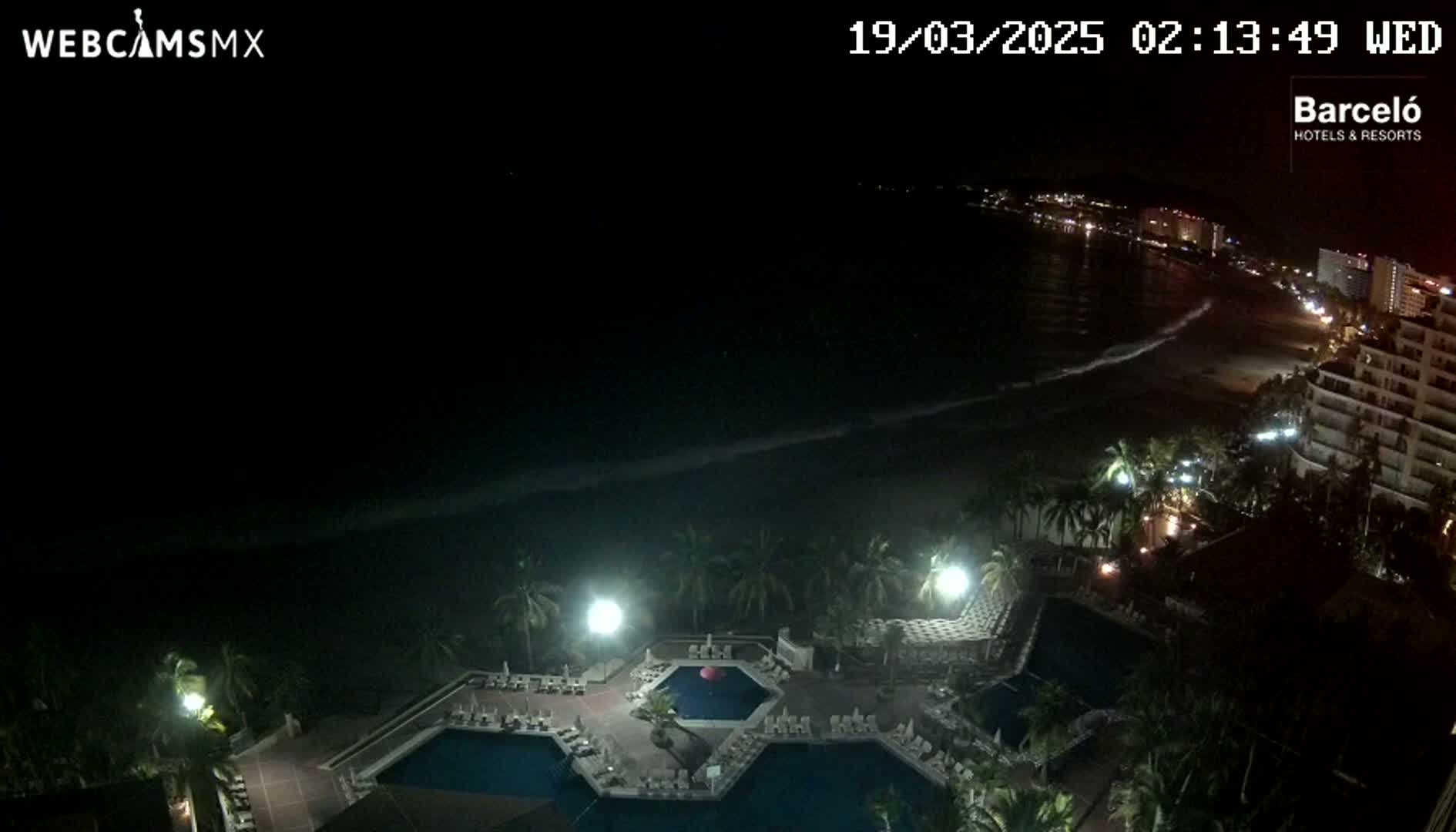 A nighttime view of a resort's pool area, illuminated by artificial lights, overlooking a dark ocean with gentle waves and a dimly lit beach.