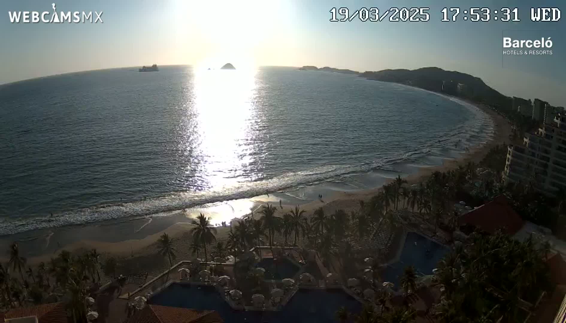 A beach with calm waves at sunset is overlooked by a resort with multiple pools, palm trees, and buildings.