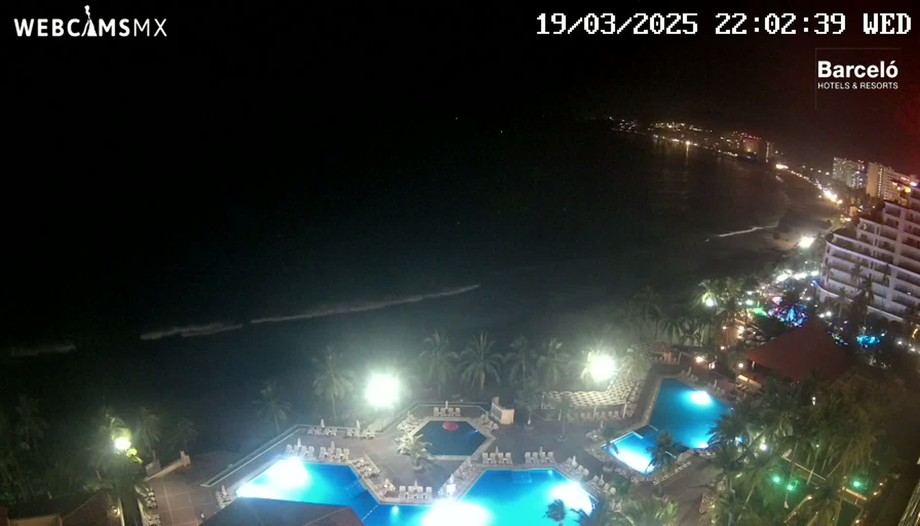 A nighttime aerial view shows a resort with illuminated pools and palm trees overlooking a dark ocean with small waves under a clear sky.