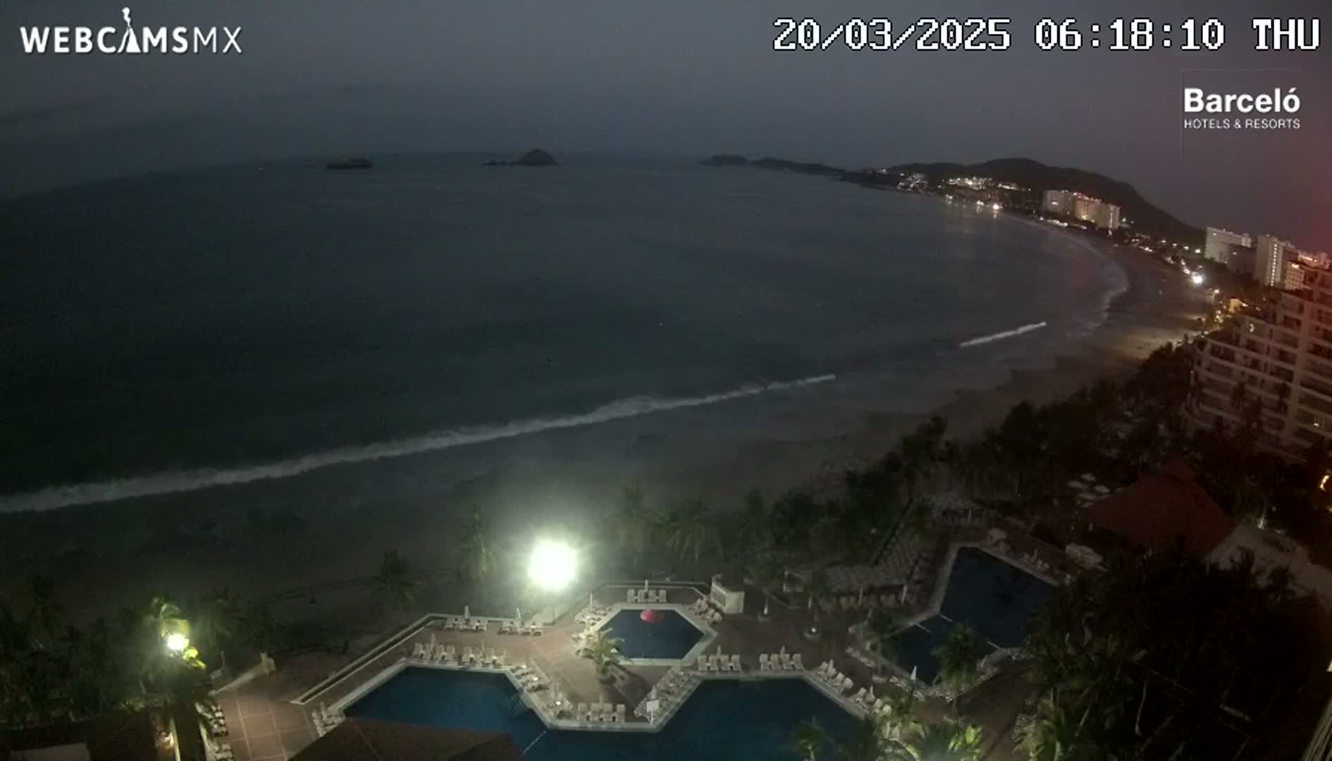 A nighttime, high-angle view shows a resort with multiple pools overlooking a dark ocean with gentle waves and a dimly lit beach.