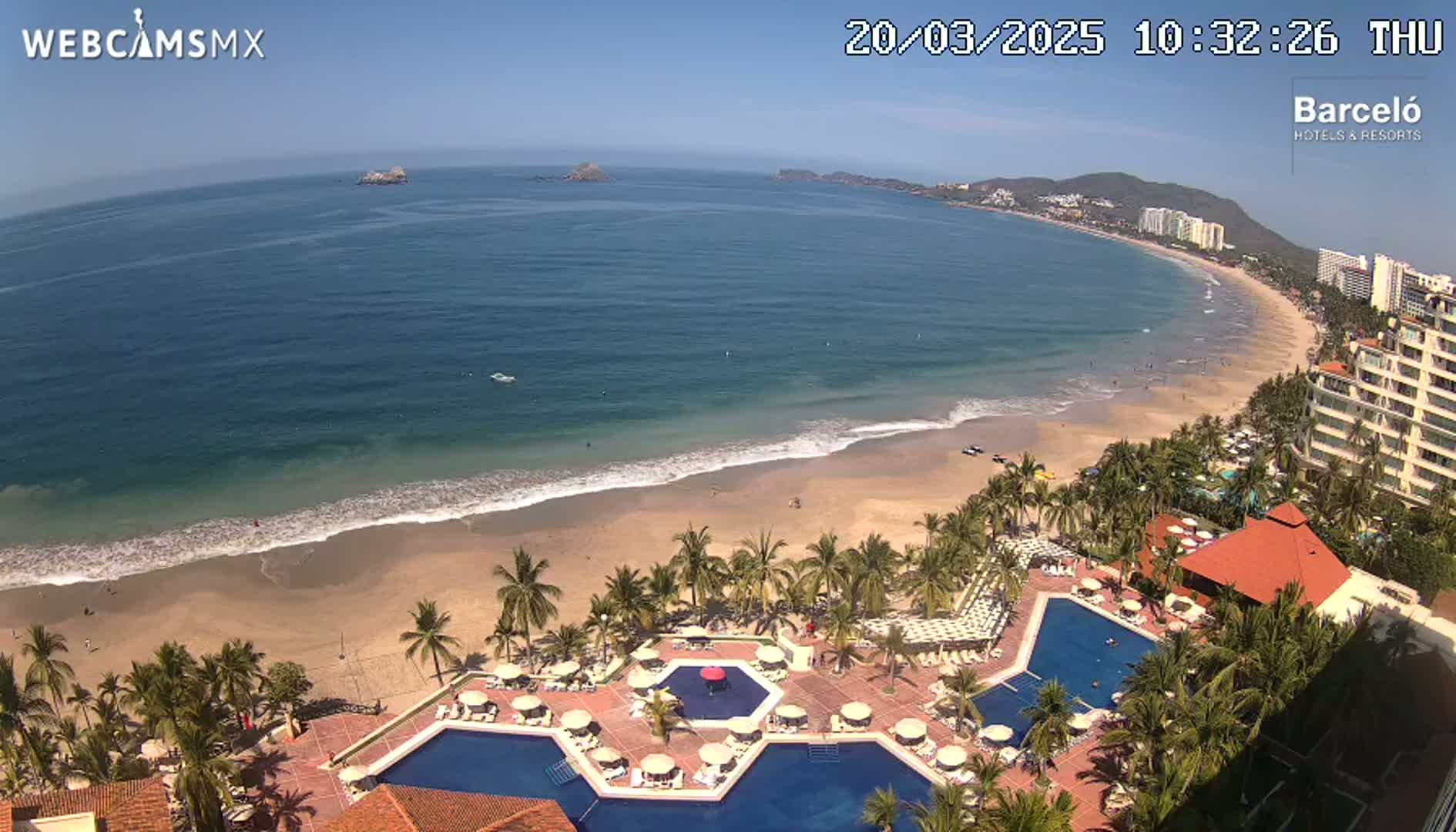 An aerial view shows a resort with multiple pools overlooking a sandy beach and calm ocean under a clear blue sky.