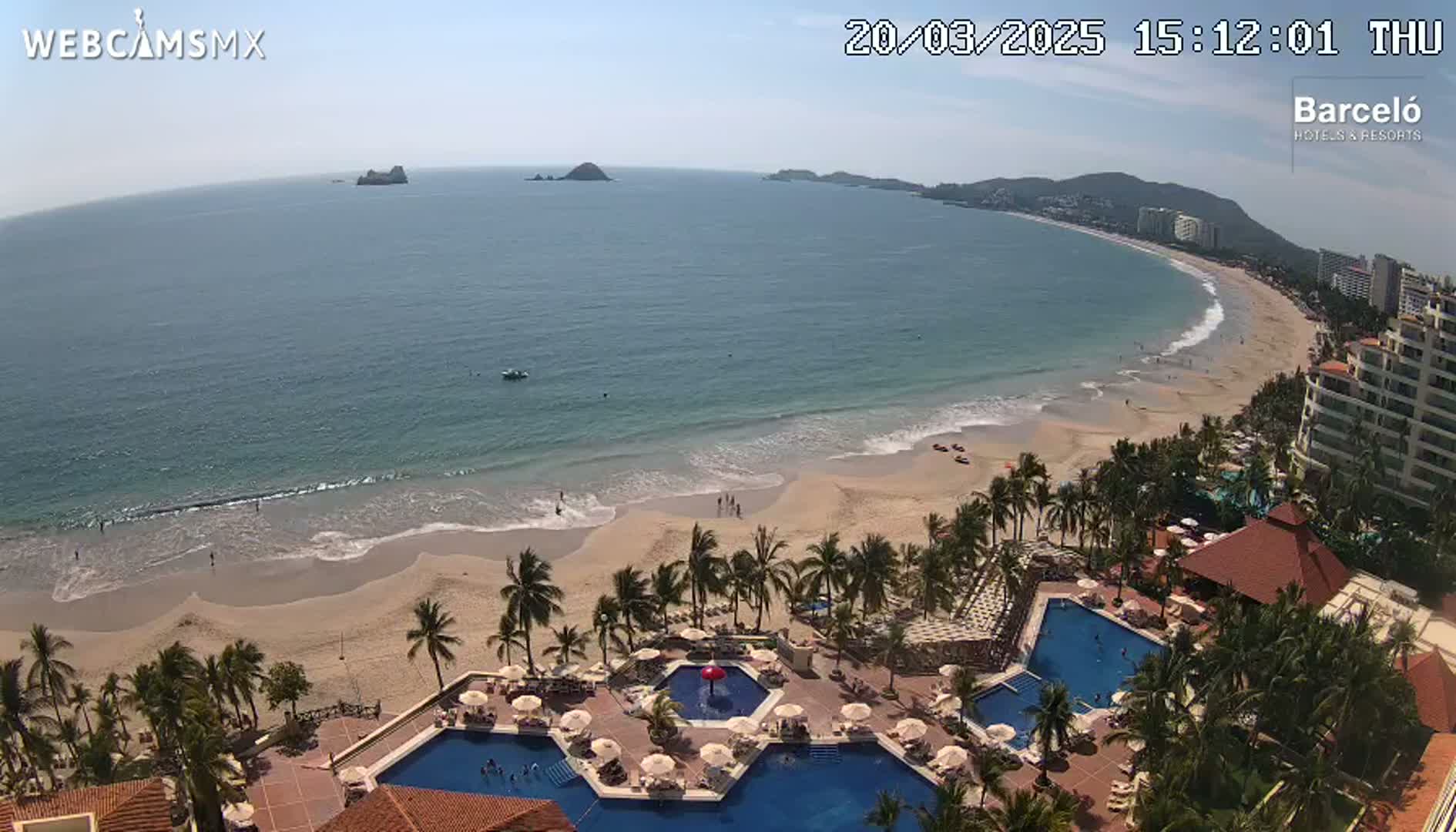 An aerial view shows a resort with multiple pools overlooking a sandy beach and calm ocean under a sunny sky.