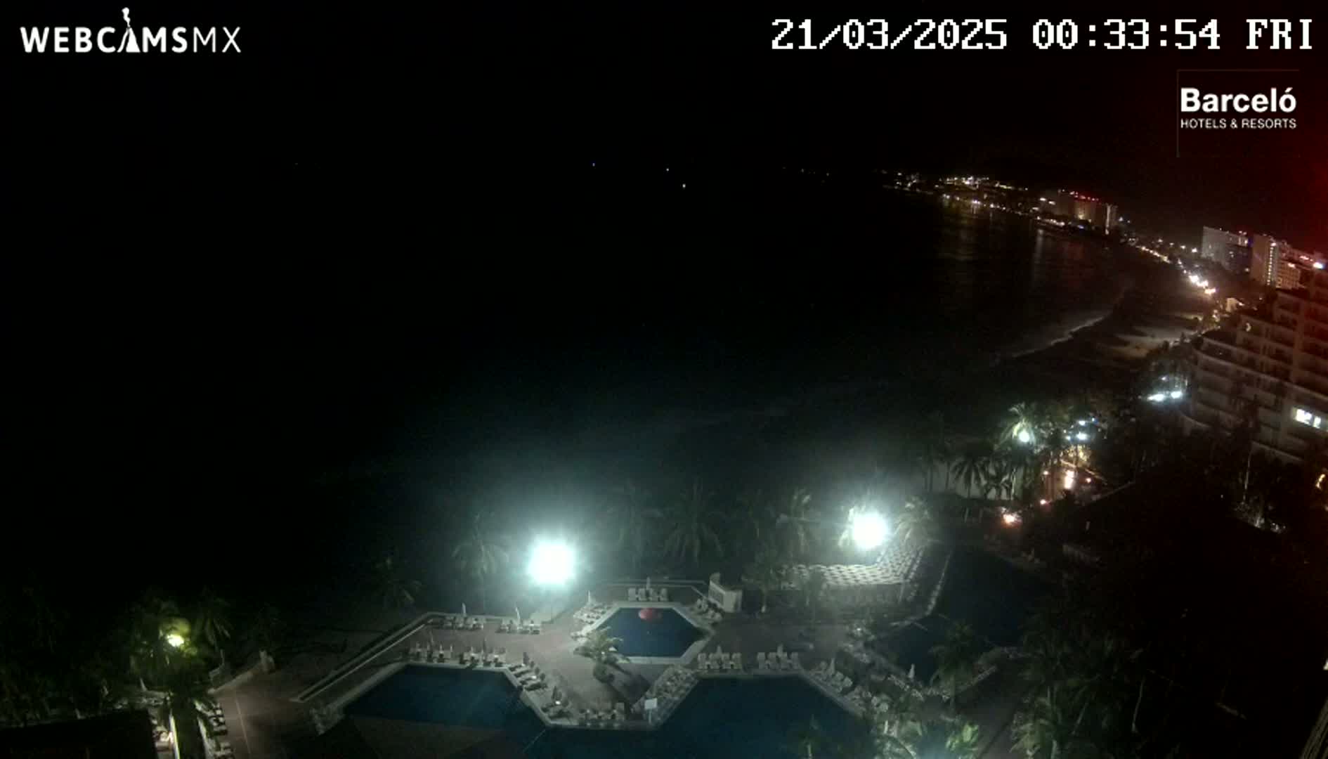 A nighttime aerial view shows a resort pool area with surrounding buildings and a dark ocean with a distant, dimly lit shoreline.