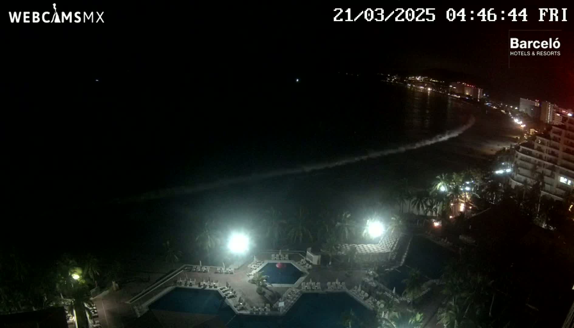 A nighttime view of a resort with illuminated pools, overlooking a dark ocean with gentle waves and a dimly lit beach and coastline.