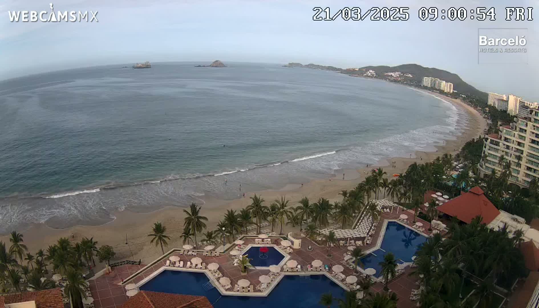 An aerial view shows a resort with multiple pools overlooking a sandy beach and calm ocean under an overcast sky.