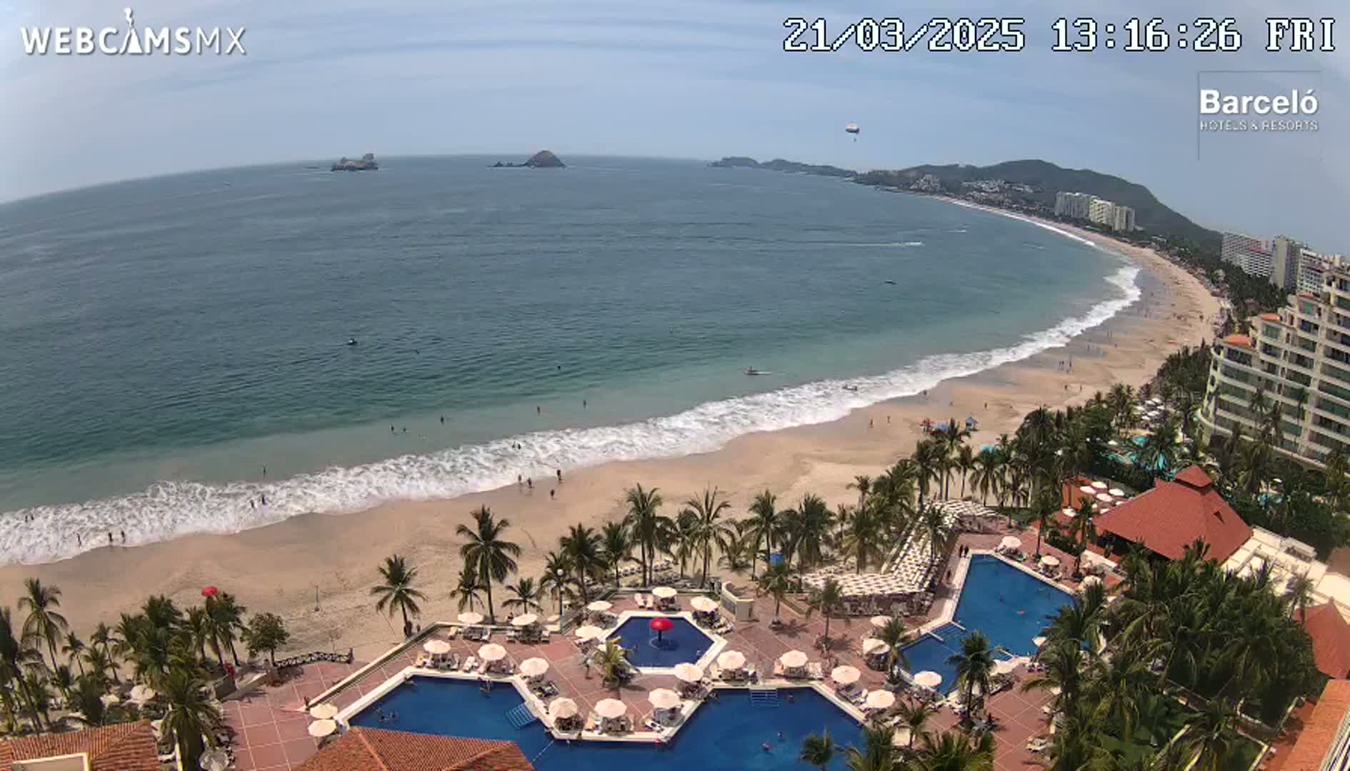 An aerial view shows a resort with multiple pools overlooking a sandy beach and calm ocean under a sunny sky.