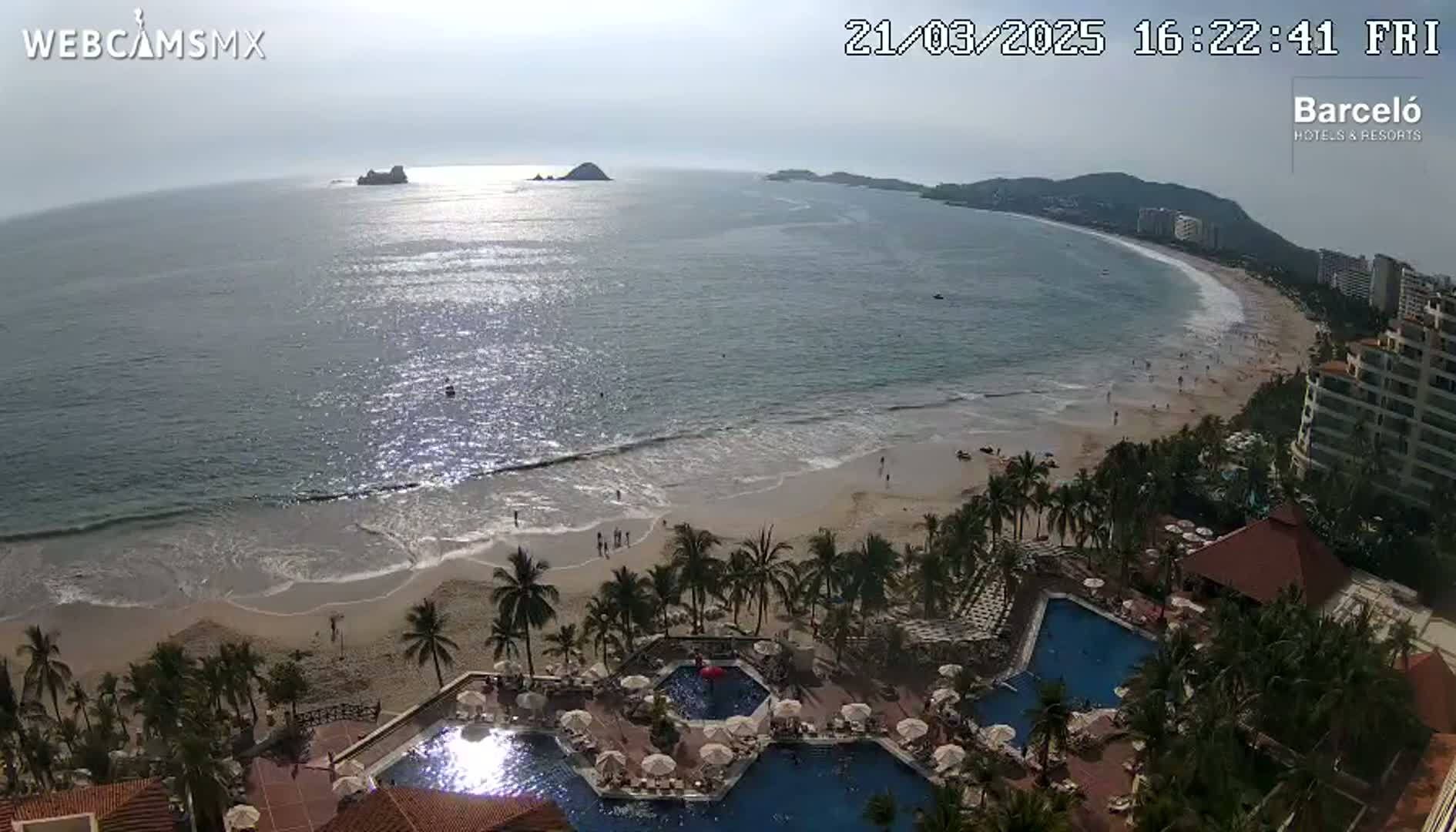 An aerial view shows a beach with calm, sunlit water, palm trees, and a resort with multiple pools.