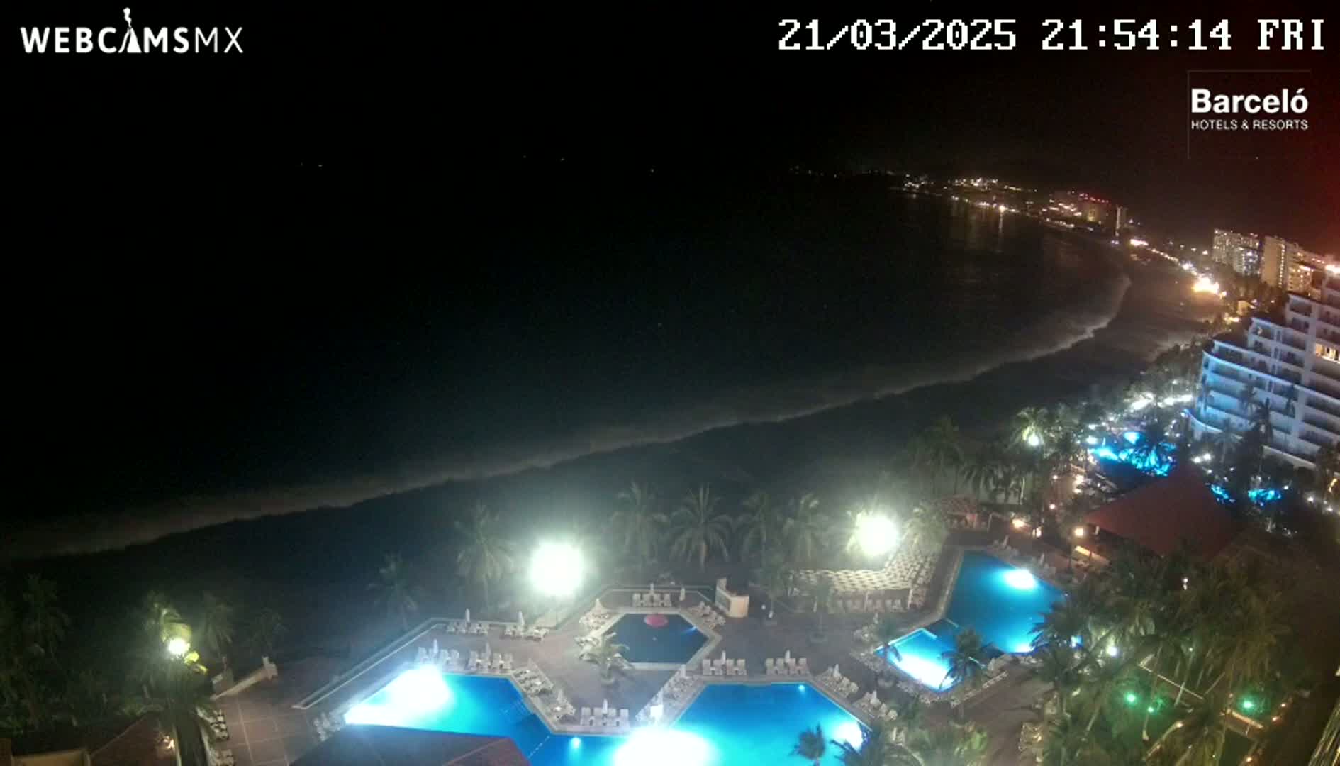 A nighttime view of a resort with illuminated pools, overlooking a dark ocean with waves under a clear sky.