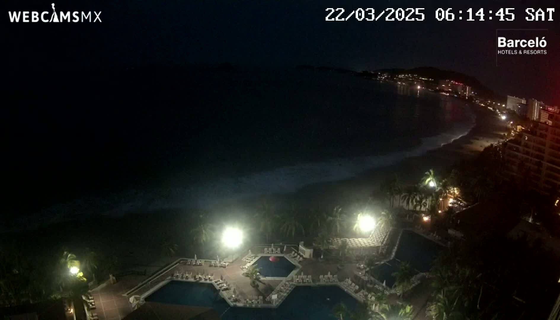 A nighttime view of a resort complex with multiple pools, illuminated by bright lights, overlooking a dark ocean with gentle waves and a dimly lit beach and shoreline.