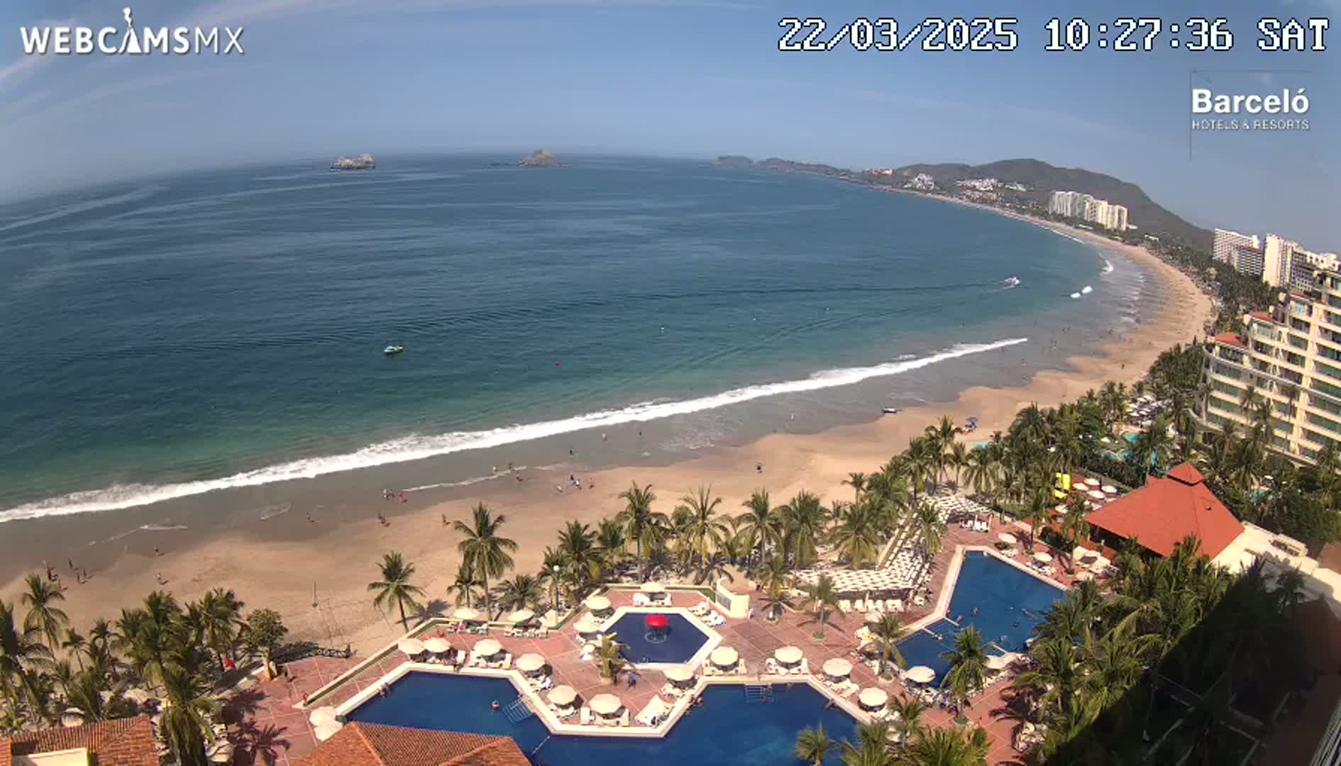 An aerial view shows a resort with multiple pools overlooking a sandy beach and calm ocean under a sunny sky.