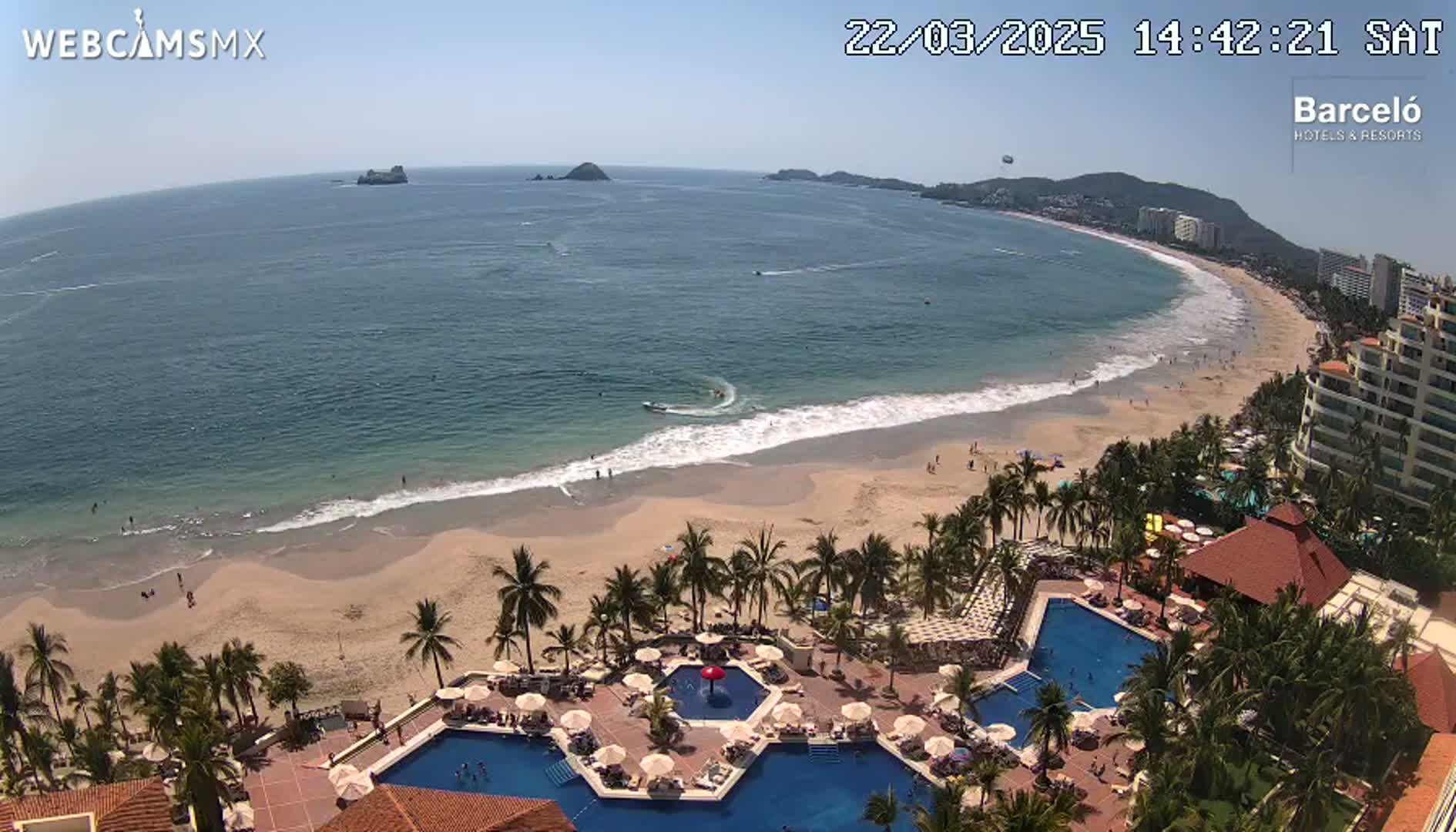 An aerial view shows a sandy beach with calm ocean waves, under a clear sunny sky, next to a resort with multiple pools and palm trees.