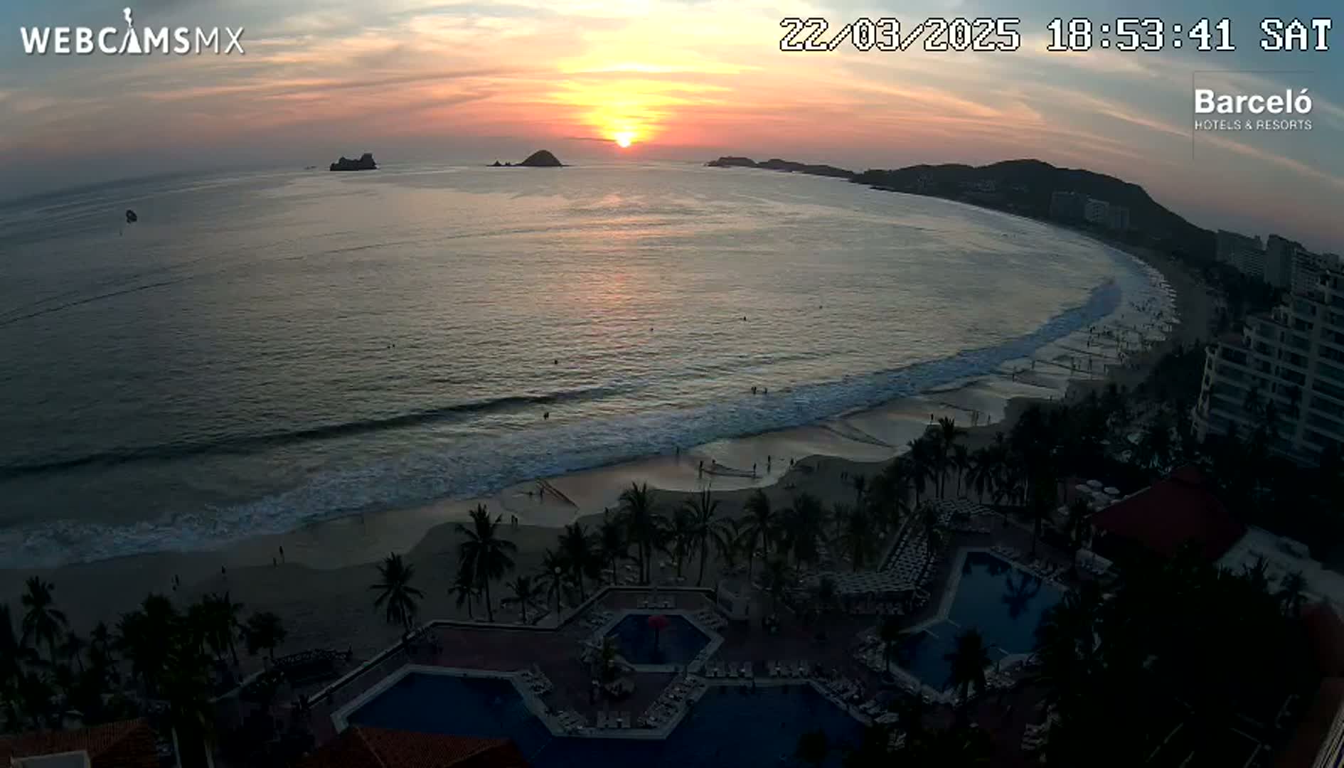 An ocean beach at sunset, with a resort and several swimming pools visible along the shore.