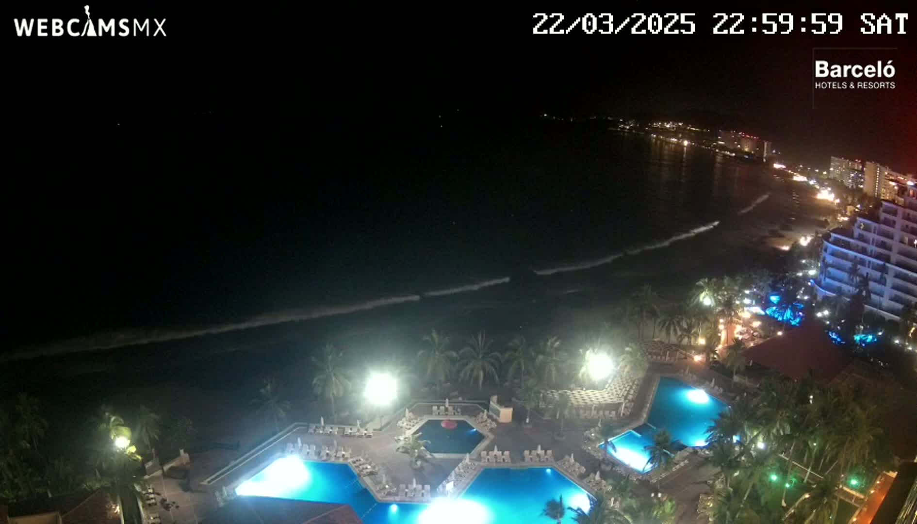 A nighttime aerial view shows a resort with illuminated pools next to a dark ocean with gentle waves under a clear, starless sky.