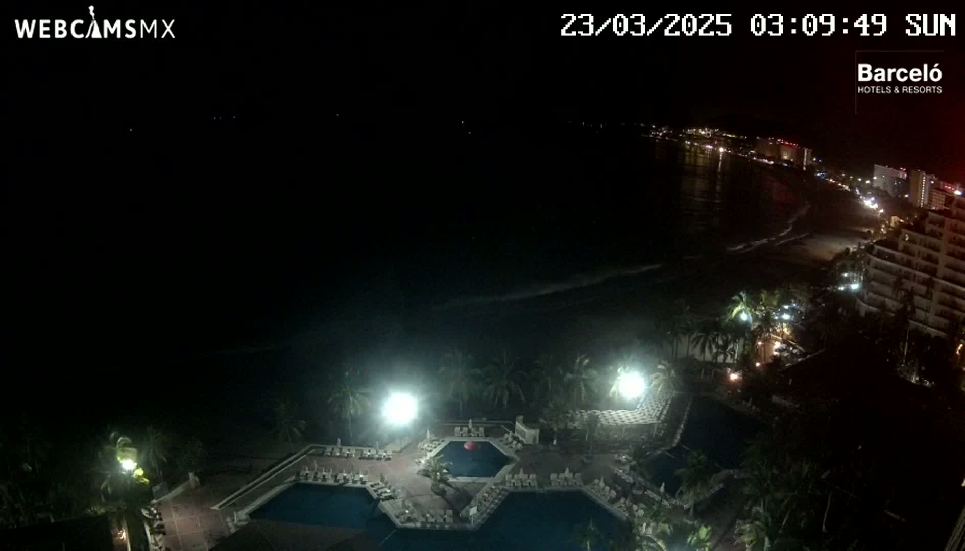 A nighttime view of a resort's pool area and a dark ocean with a city's lights visible in the distance.