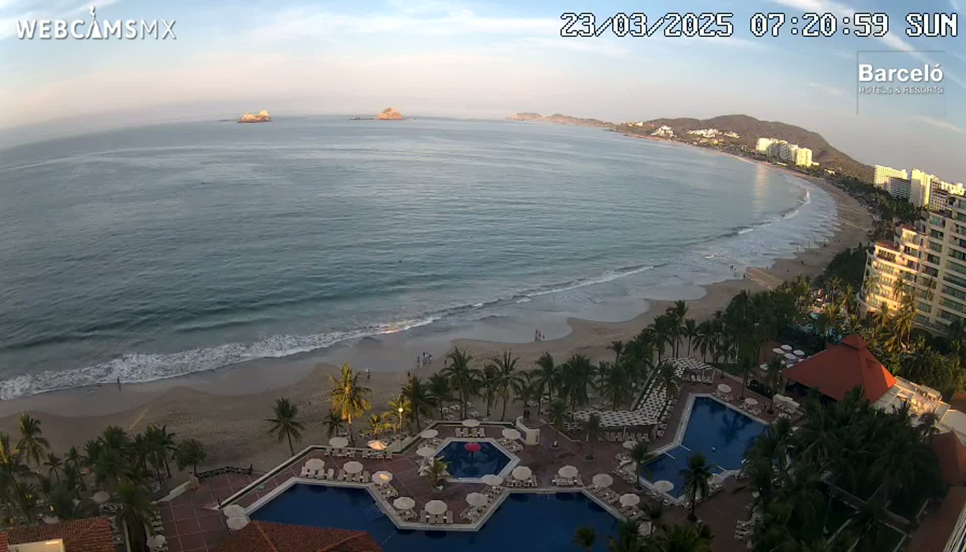 An aerial view shows a resort with multiple pools next to a sandy beach and ocean under a partly cloudy sunrise.