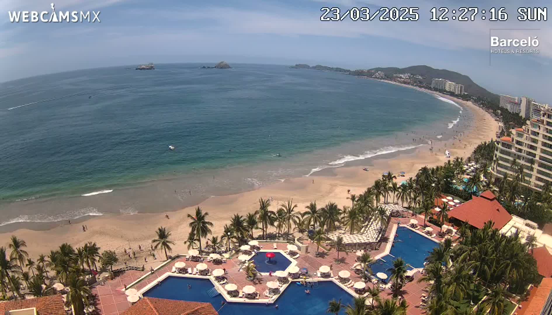 An aerial view shows a resort with multiple pools overlooking a sandy beach and calm ocean on a sunny day.