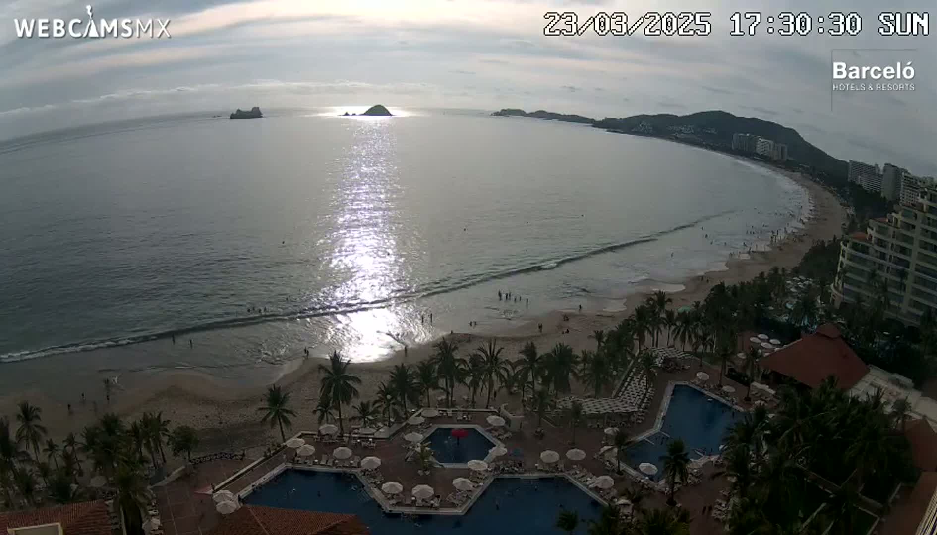 An aerial view shows a beach with calm waves, palm trees, a resort with multiple pools, and a small island in the distance under a partly cloudy sky.