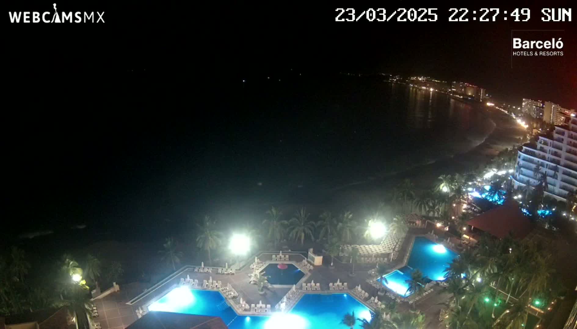 A nighttime view of a resort with illuminated pools overlooking a dark ocean and a dimly lit beach with buildings in the distance.