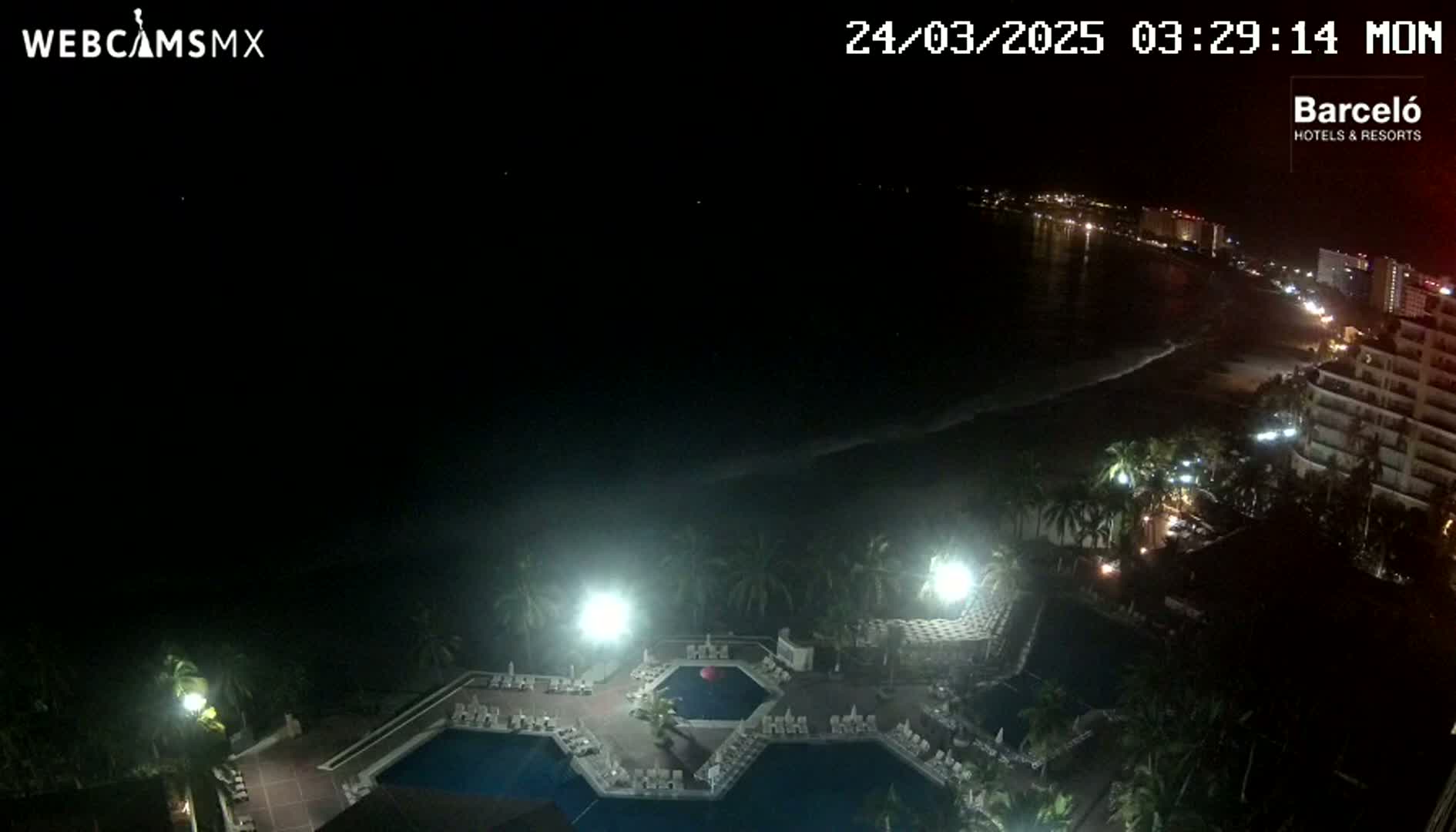 A nighttime aerial view shows a resort pool area illuminated by bright lights, with a dark ocean and a dimly lit shoreline visible in the background.