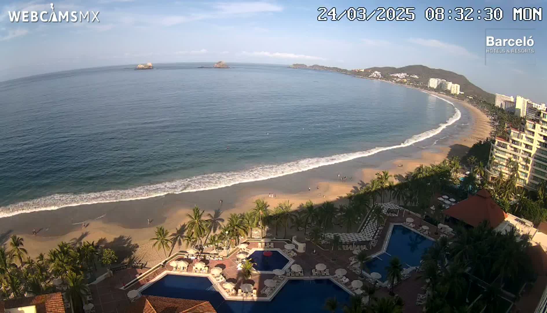 An expansive sandy beach with gentle waves laps a tranquil ocean under a mostly clear sky, adjacent to a resort with multiple pools and palm trees.