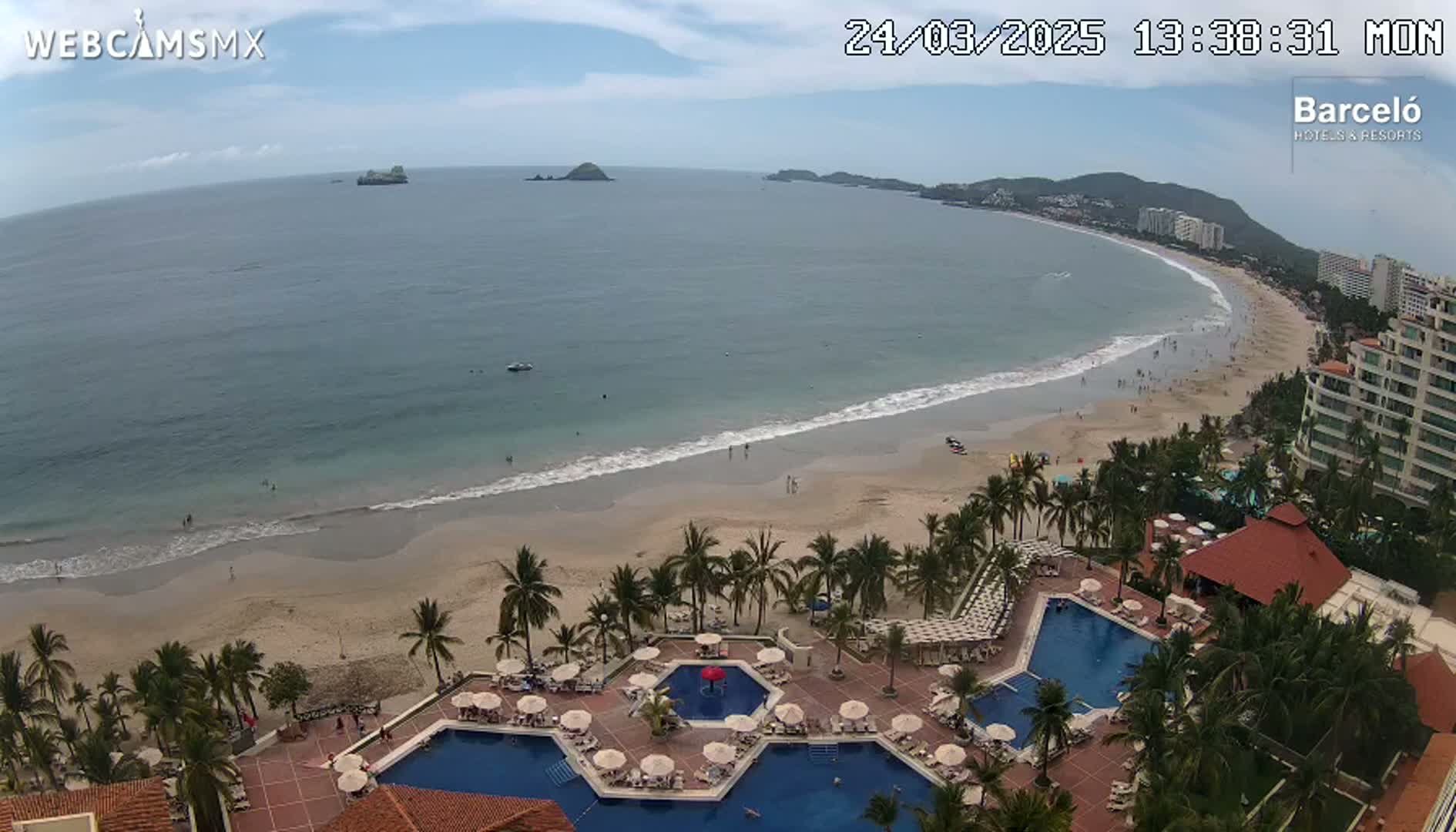 An aerial view shows a resort with multiple pools overlooking a sandy beach and calm ocean under a partly cloudy sky.
