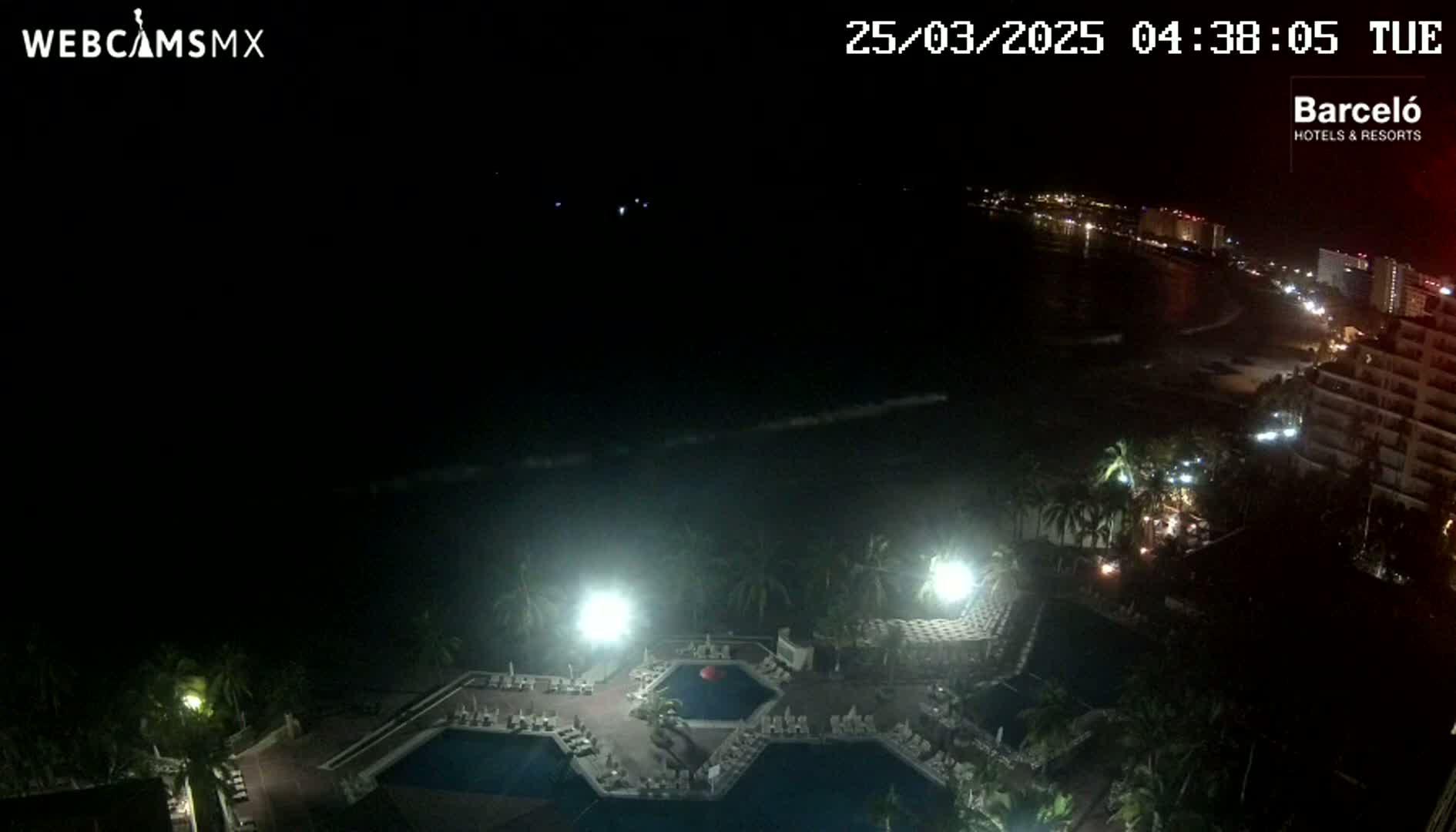 A nighttime aerial view shows a resort area with pools and palm trees illuminated by lights, overlooking a dark ocean and a dimly lit coastline.