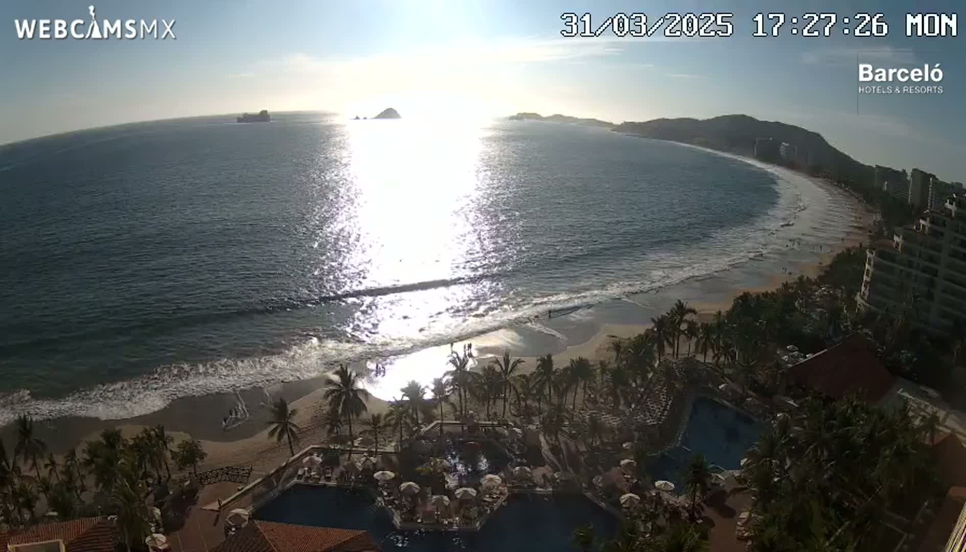 An aerial view shows a beach with calm waves under a sunny sky, palm trees lining the shore, and a resort with multiple pools nestled amongst the vegetation.