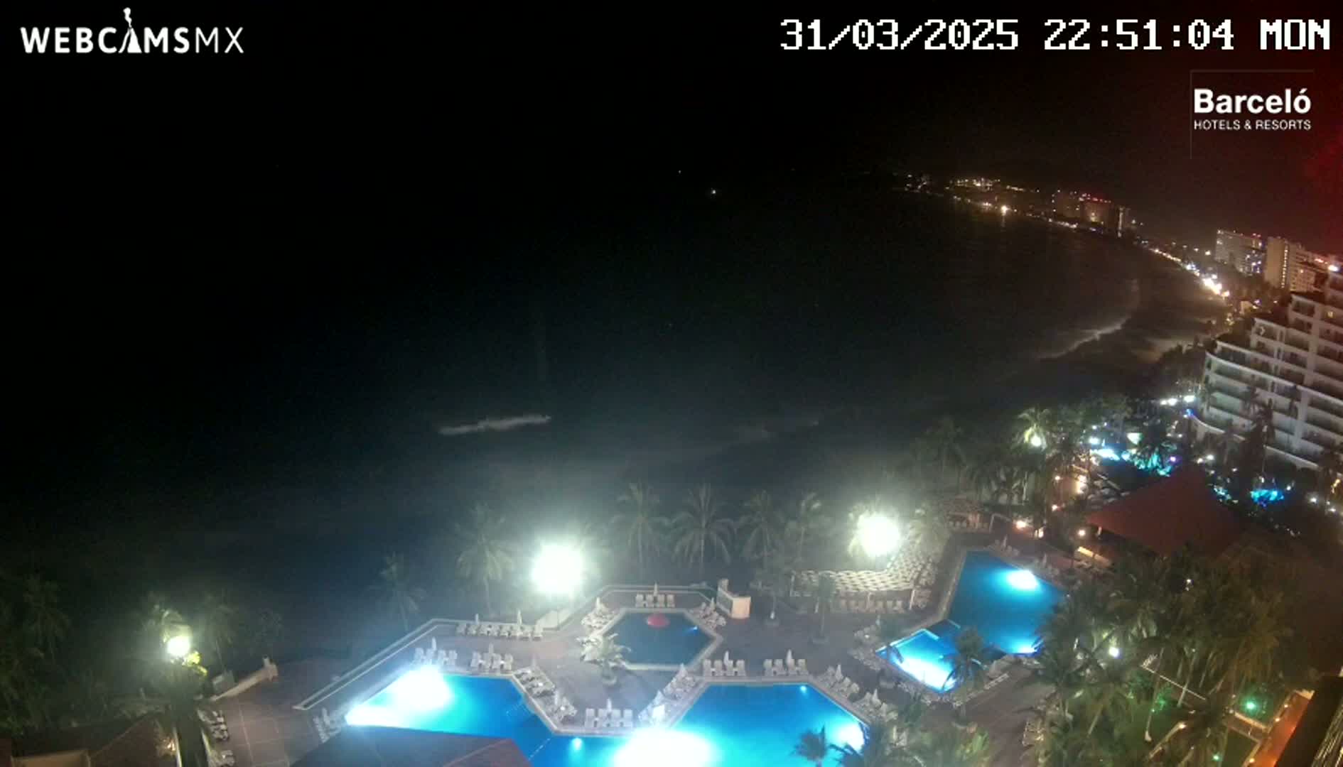 A nighttime view of a resort's illuminated pools and surrounding coastline, with a dark ocean visible under a clear sky.