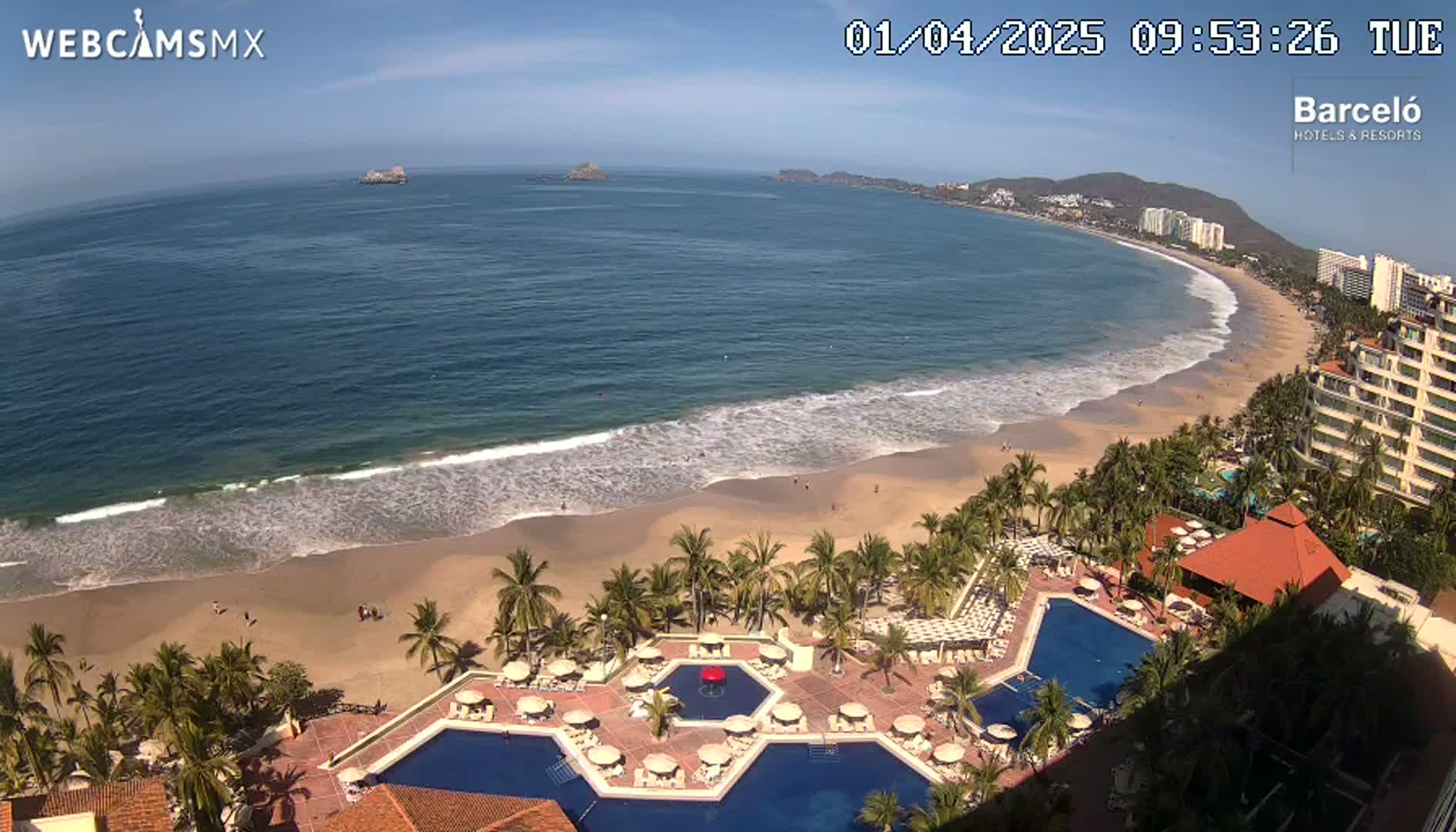 An aerial view shows a beach with gentle waves under a sunny sky, palm trees, and a resort with multiple pools.