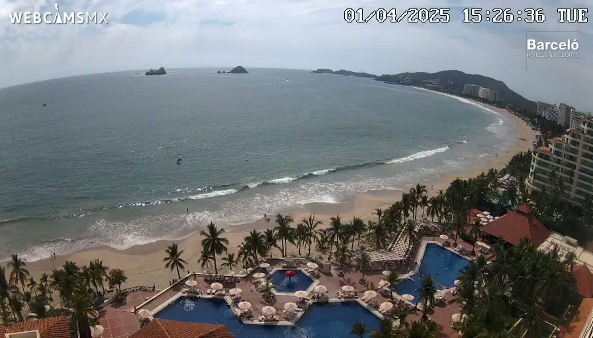 An aerial view of a resort with multiple pools overlooking a beach with gentle waves under a partly cloudy sky.