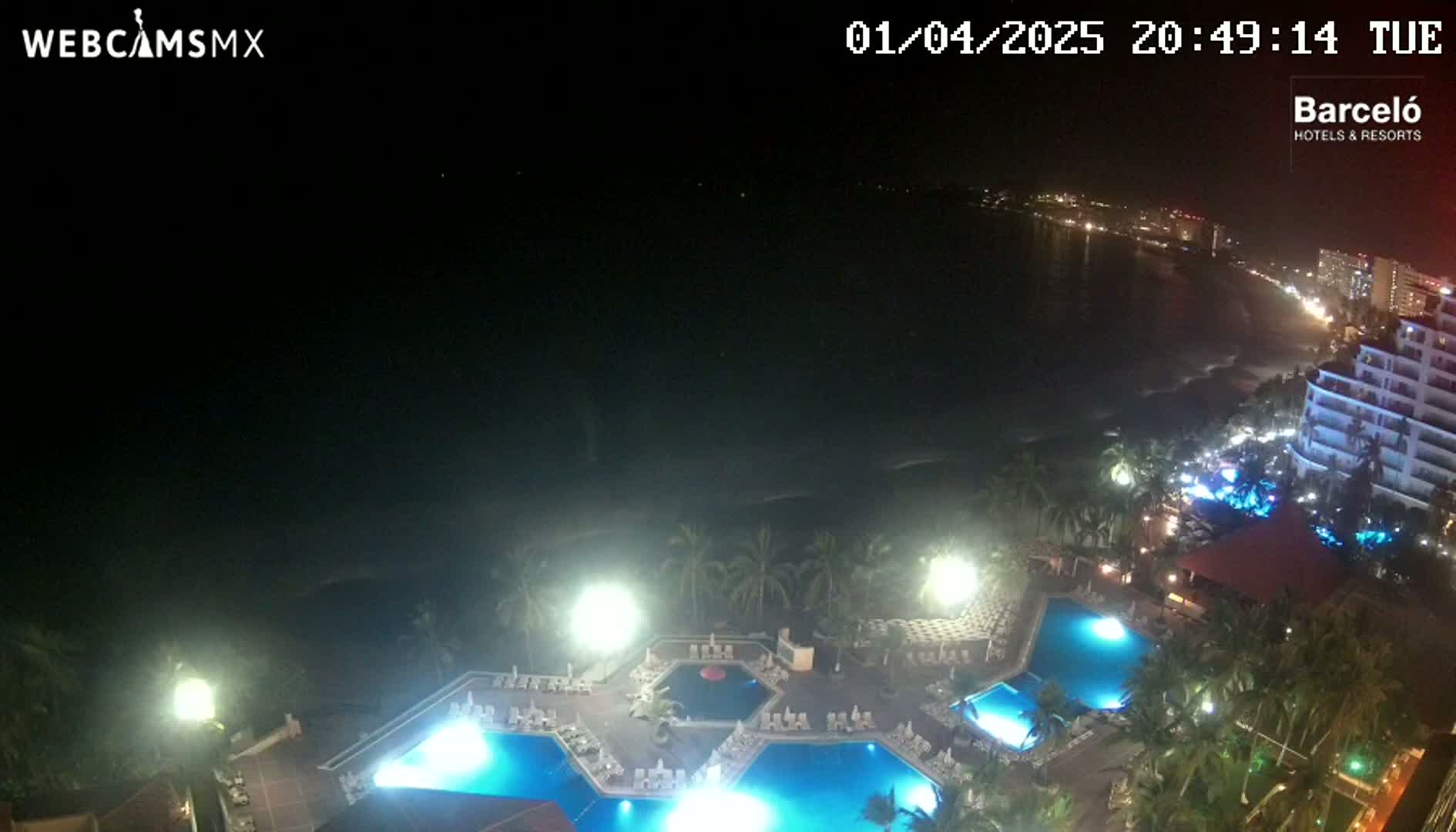 An illuminated resort at night overlooks a dark ocean with a city skyline visible in the distance under a clear sky.