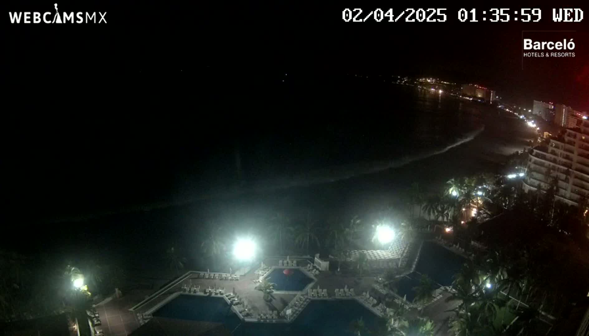 A nighttime aerial view shows a resort with multiple pools and surrounding vegetation, overlooking a dark ocean with a gently lit shoreline in the distance.
