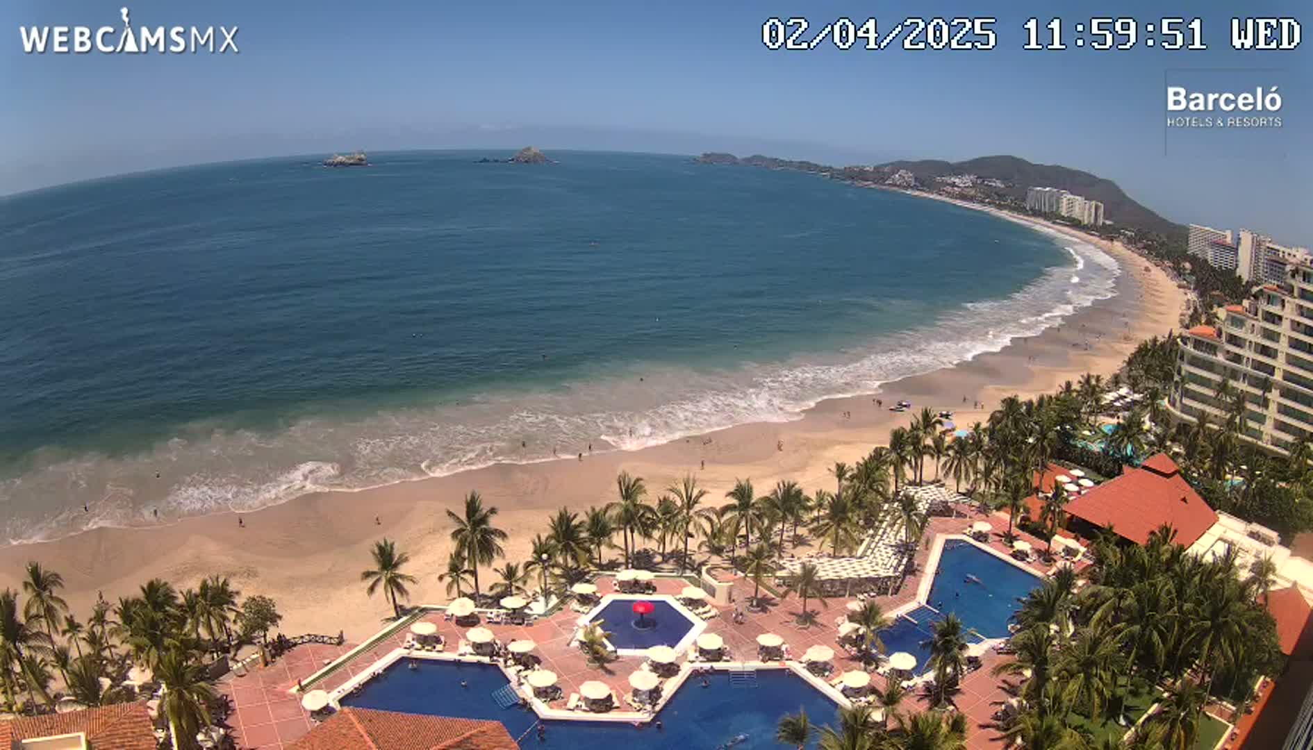An aerial view shows a resort with multiple pools next to a sandy beach and ocean under a clear blue sky.