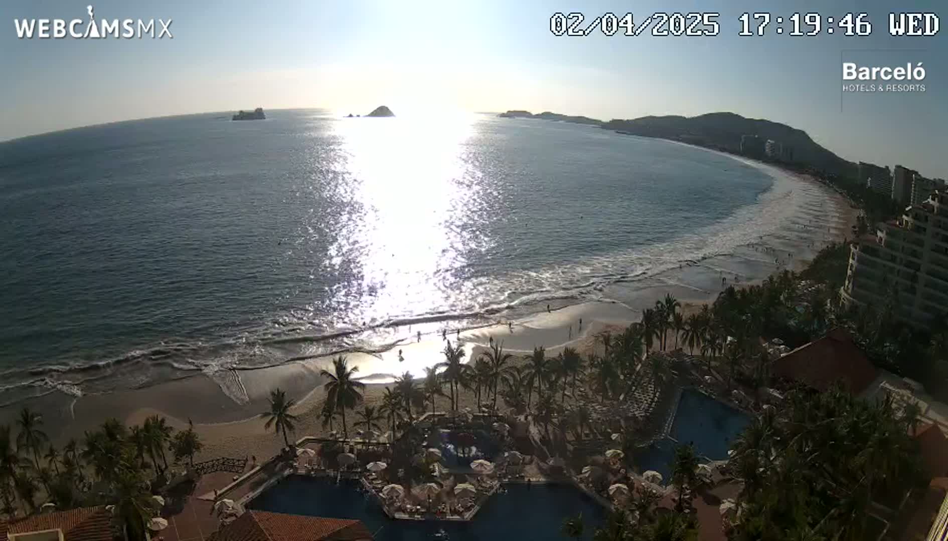 An ocean beach scene at sunset shows a sandy shore, palm trees, resort pools, and buildings, with calm waves lapping the sand under a clear sky.