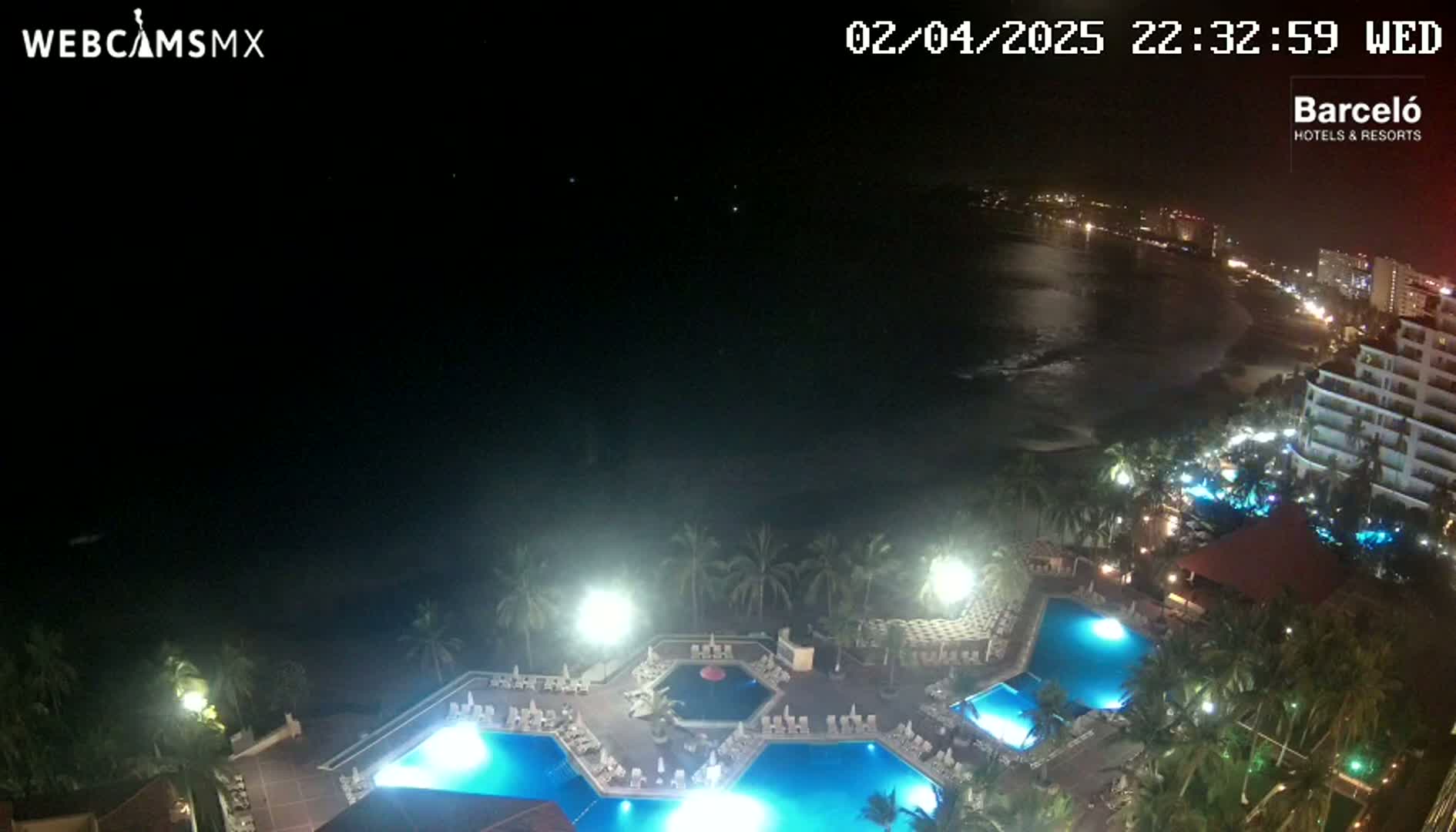 A nighttime aerial view shows a resort with illuminated pools and palm trees, overlooking a dark ocean and a city skyline.