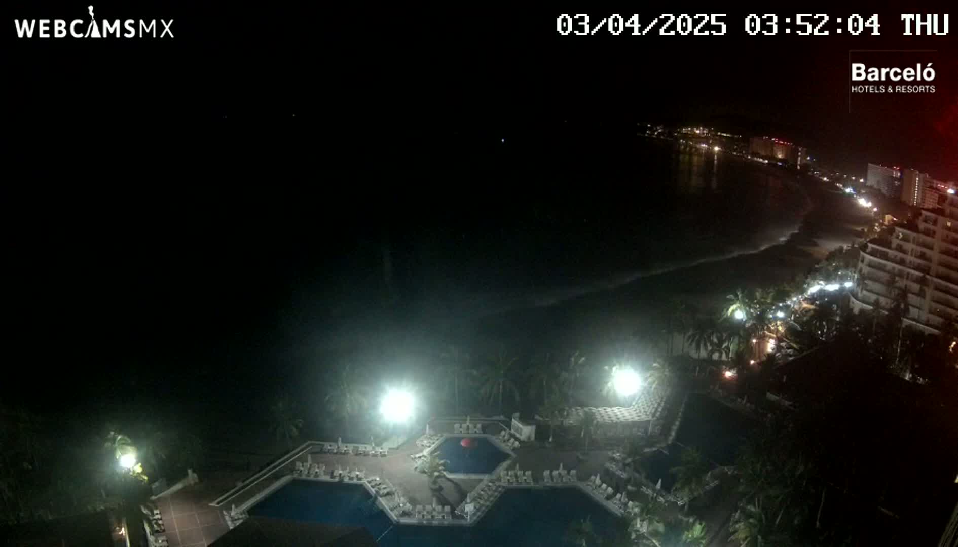 A nighttime aerial view shows a resort pool area with surrounding buildings and a dark ocean with a distant, dimly lit shoreline.