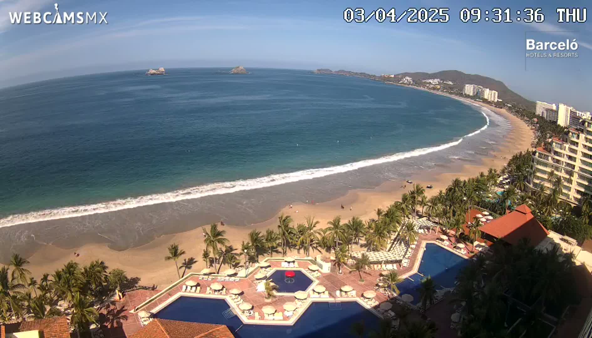 An aerial view shows a beach with gentle waves under a sunny sky, next to a resort with palm trees and multiple swimming pools.