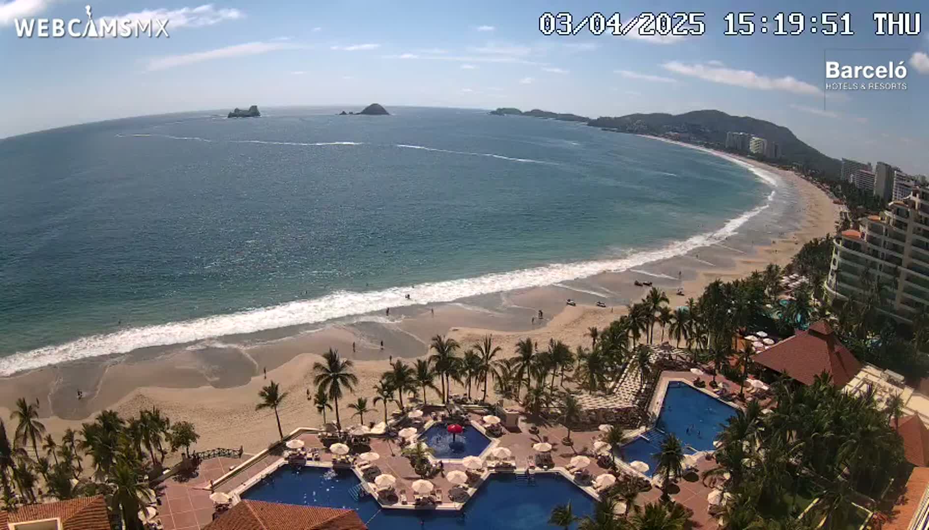 An aerial view shows a resort with multiple pools overlooking a sandy beach and calm ocean under a mostly sunny sky.