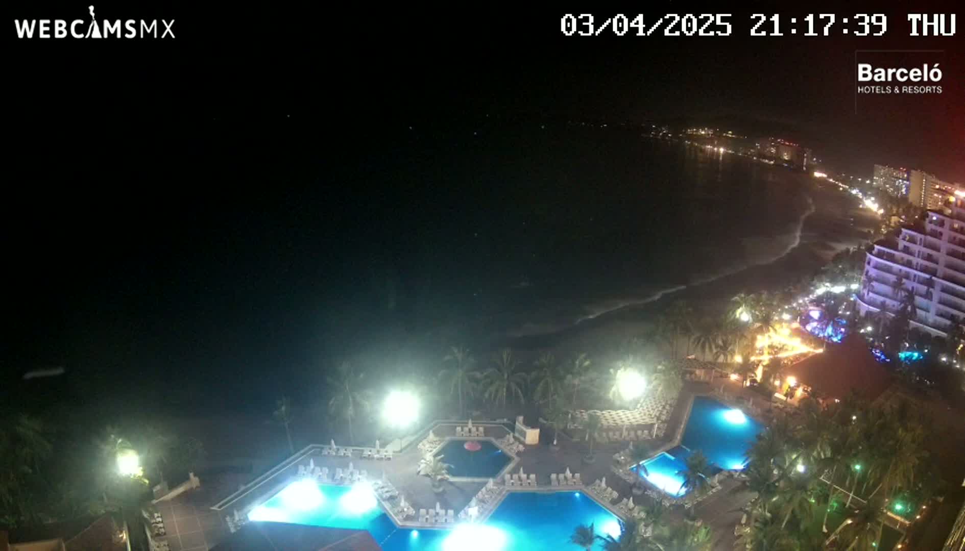 A nighttime view of a resort with multiple illuminated pools overlooking a dark ocean with gently lapping waves on a beach lined with lights.