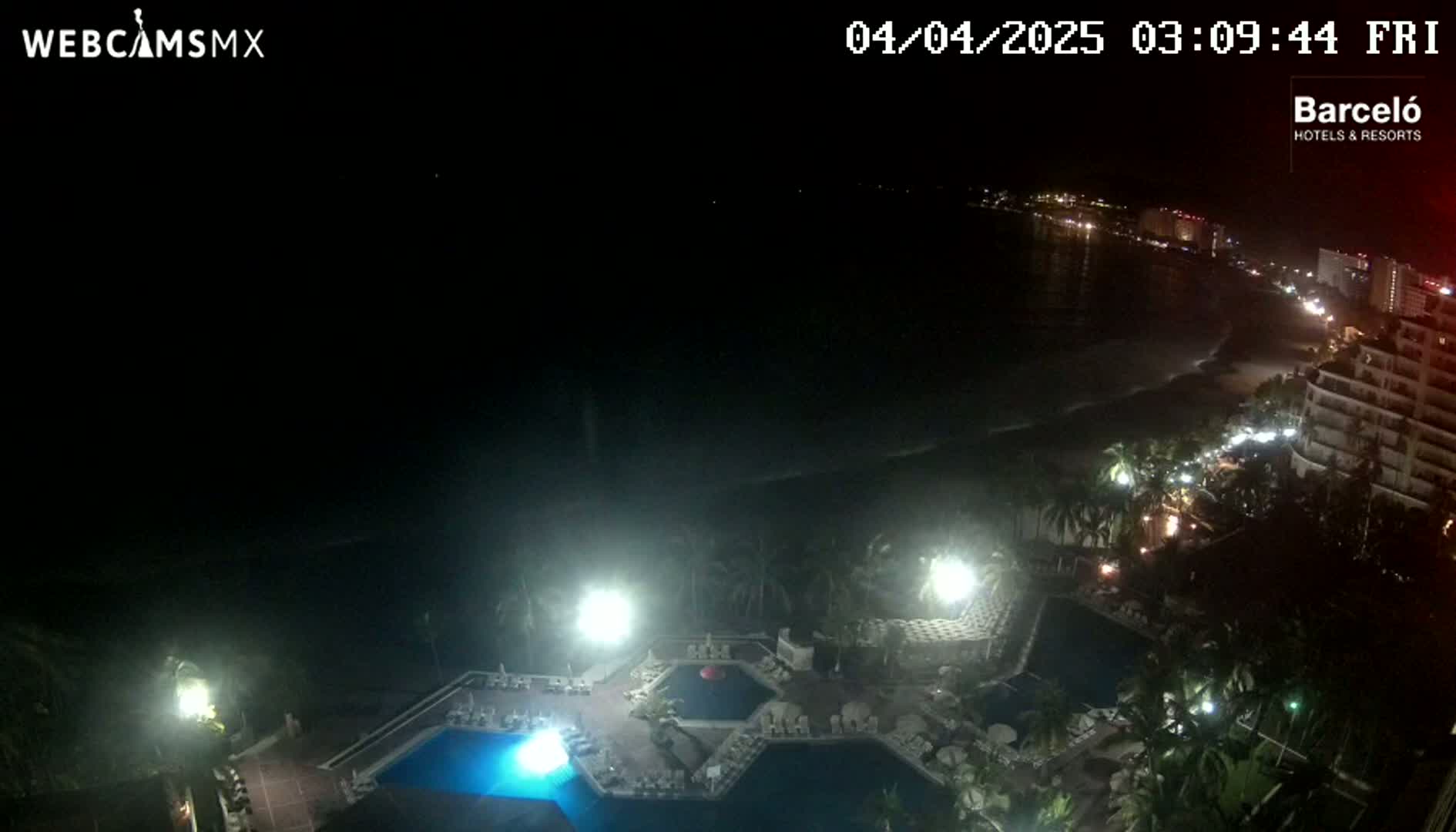 An aerial nighttime view shows a resort area with illuminated pools and palm trees, overlooking a dark ocean and a dimly lit beach.