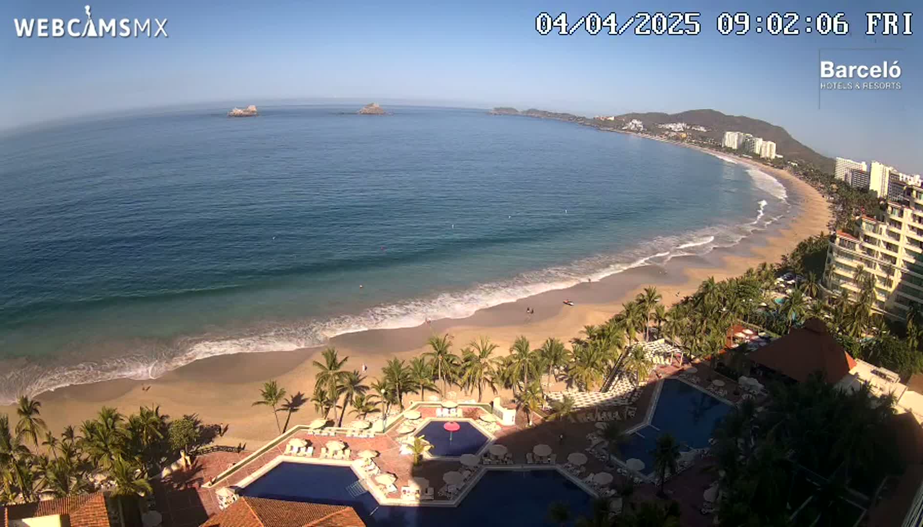 An aerial view shows a resort with multiple pools overlooking a sandy beach and a calm ocean on a sunny day.