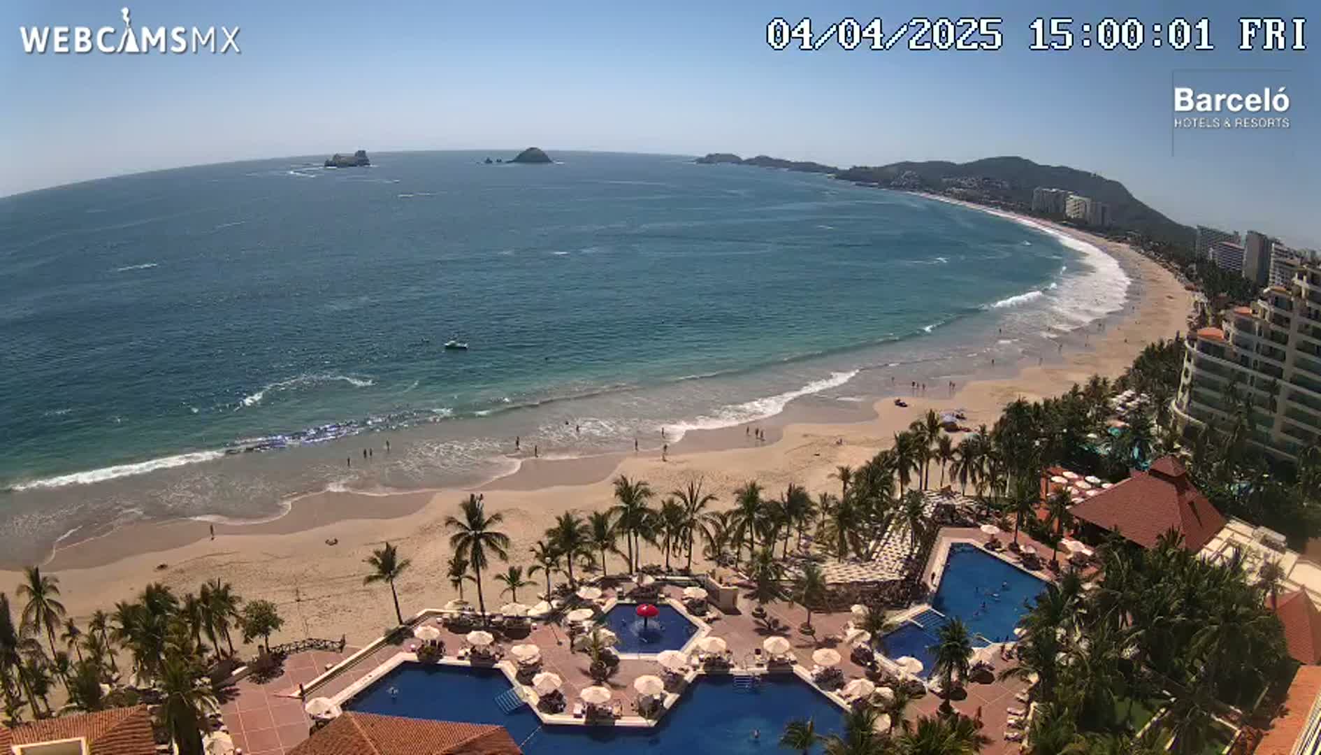 An aerial view shows a beach with palm trees and a resort with multiple pools under a sunny sky.