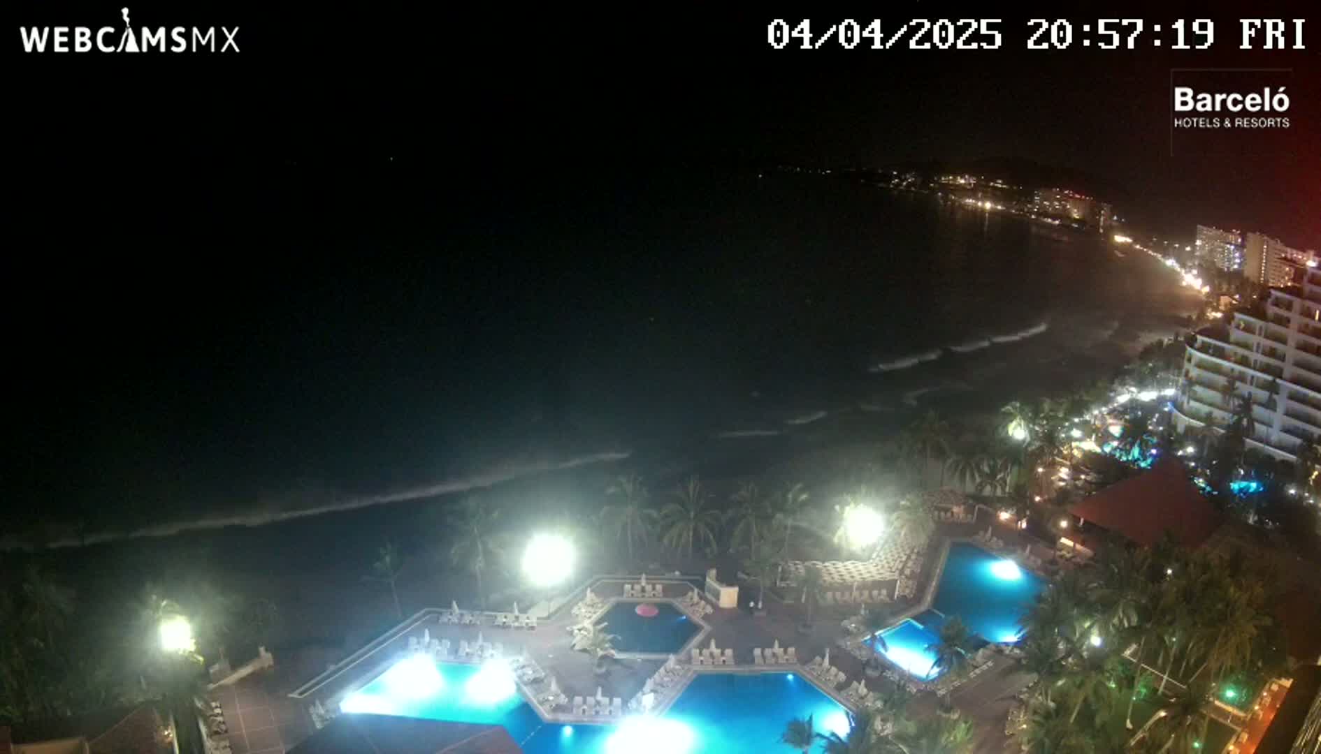 A nighttime view of a resort with multiple illuminated pools, surrounded by palm trees and overlooking a dark ocean with waves under a clear, dark sky.