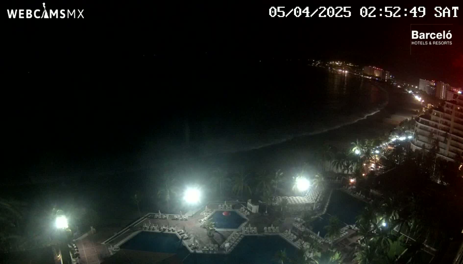A nighttime aerial view shows a resort with several illuminated pools, palm trees, and a beach with gentle waves under a clear, dark sky.