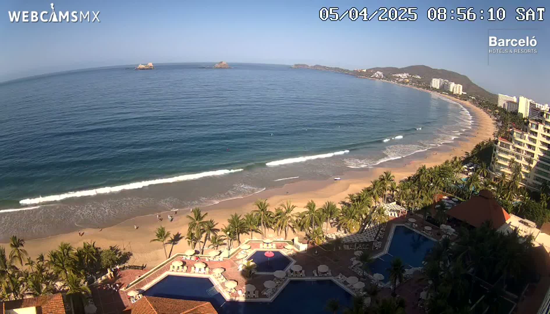 An aerial view shows a beach with calm waves lapping the shore, palm trees, and a resort with multiple pools on a sunny day.