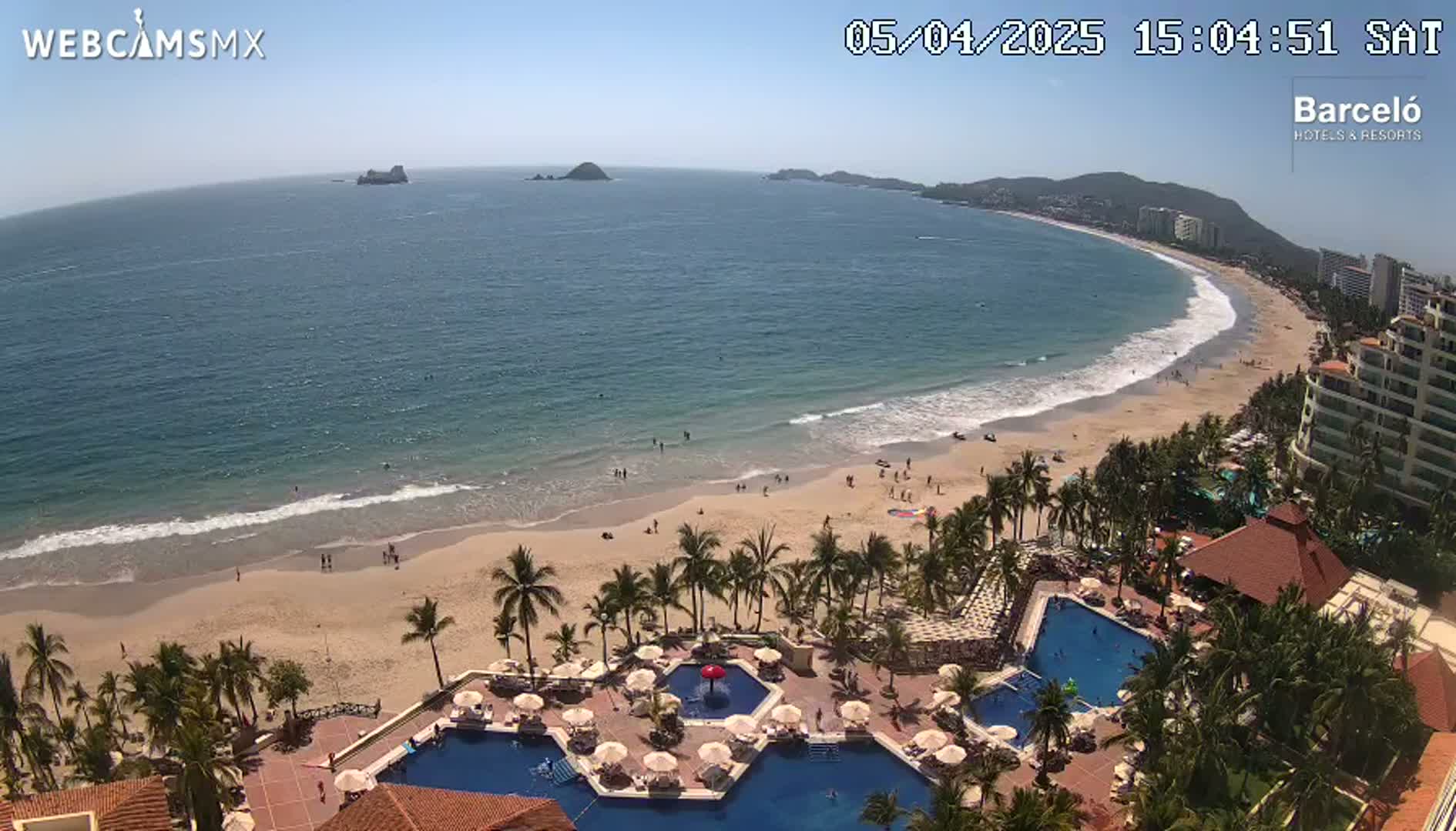 An aerial view shows a resort with multiple pools overlooking a beach with gentle waves under a clear, sunny sky.