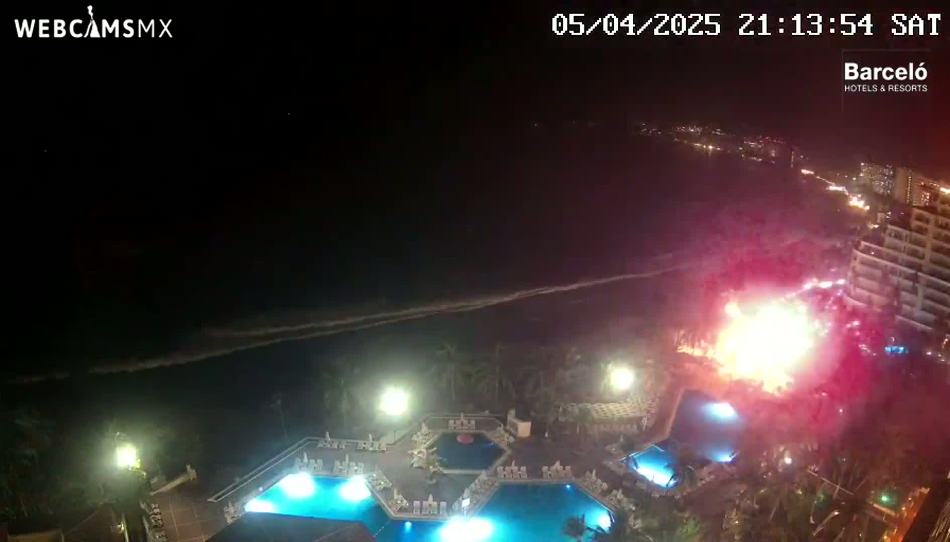 A nighttime view of a resort with multiple illuminated pools, a beach, and a fireworks display over the ocean.